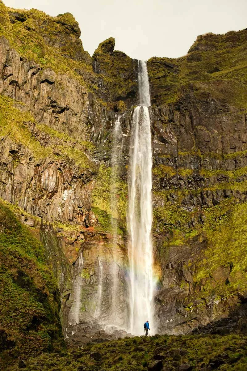 A person standing at the base of a tall waterfall cascading down a rocky, moss-covered cliff with a rainbow forming at the bottom.