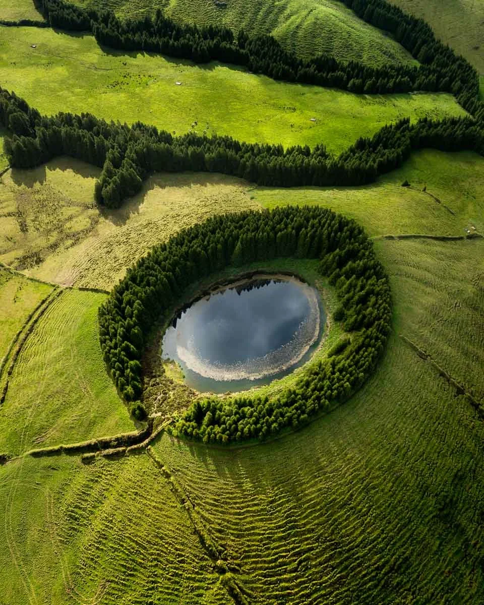 Aerial view of a circular lake surrounded by dense forest on a green landscape with fields and small trails.