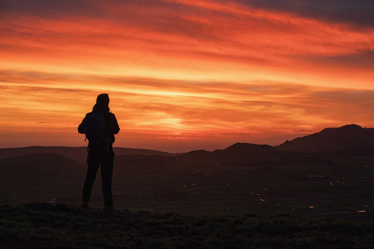 Silhouette of a person with a backpack standing on a hill at sunset, overlooking a landscape with mountains and fields.