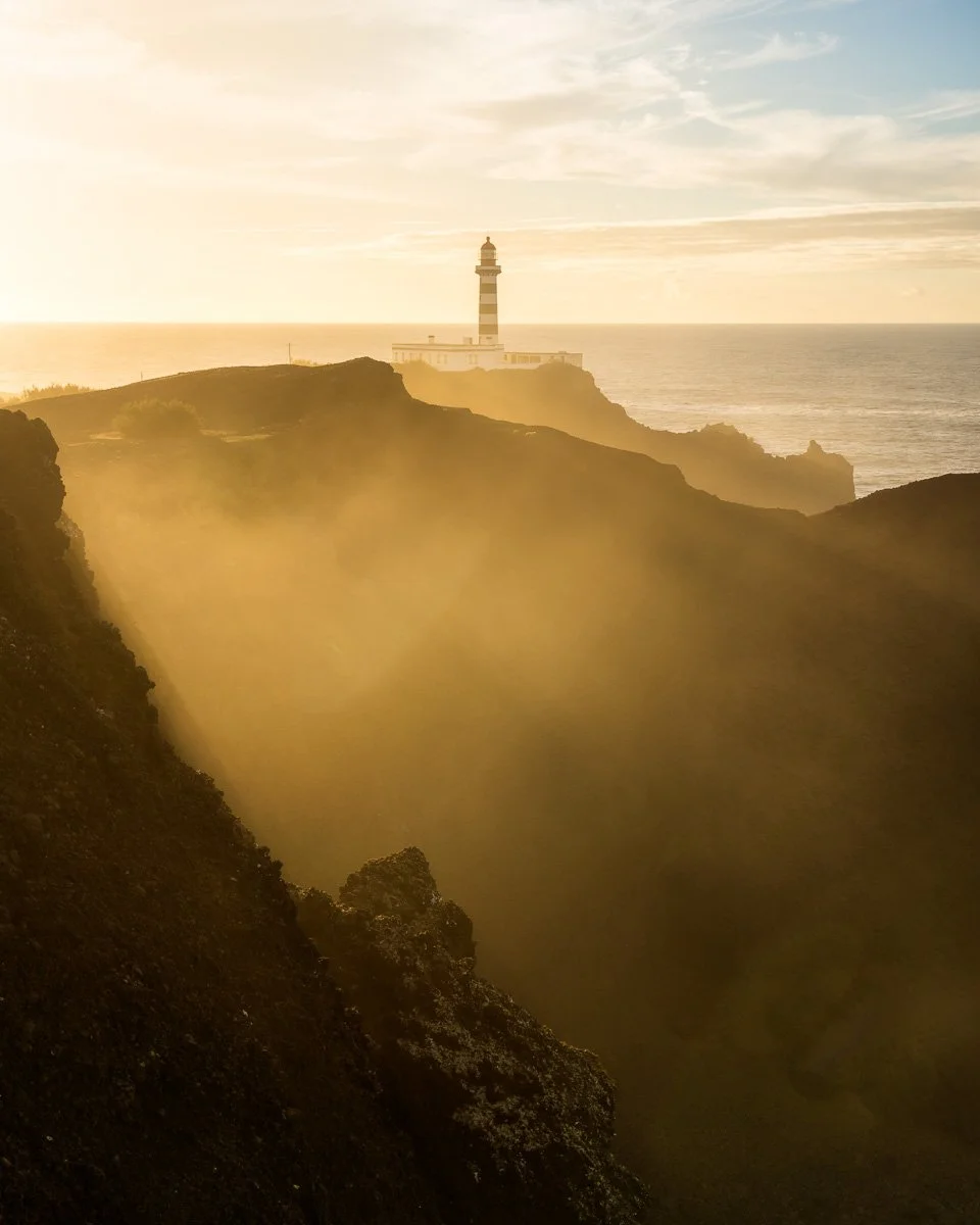 A lighthouse on a cliff overlooking the ocean during sunset, with mist and rugged terrain in the foreground.