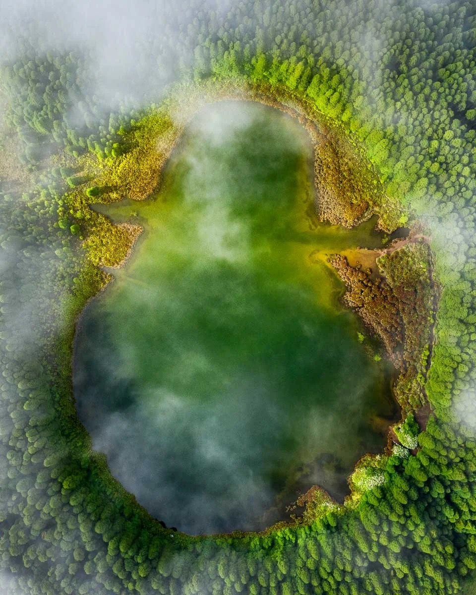 Aerial view of a green lake surrounded by dense forest with mist and clouds above.