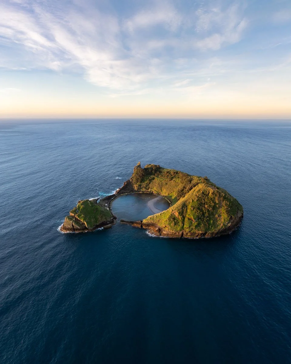 An aerial view of a small, green island with cliffs surrounding a circular, calm lagoon in the ocean, under a partly cloudy sky at sunset.