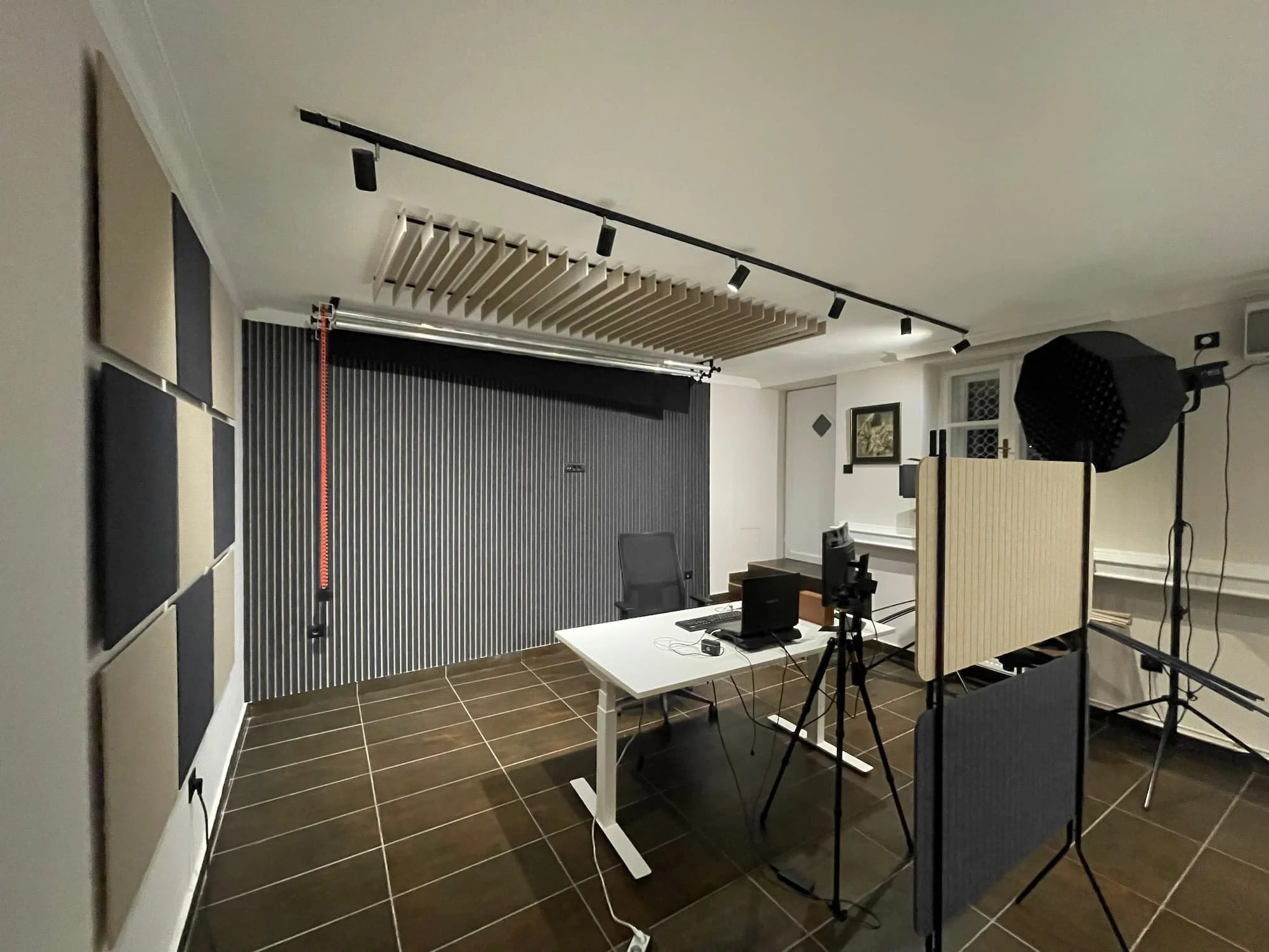 Podkast studio sa crno - bež zidom, zvučno-izolacionim akustičnim filc panelima, profesionalnom rasvetom, kamerom na stalku i belim radnim stolom sa računarskom opremom. Idealno rešenje za kontrolu zvuka i akustiku studija.