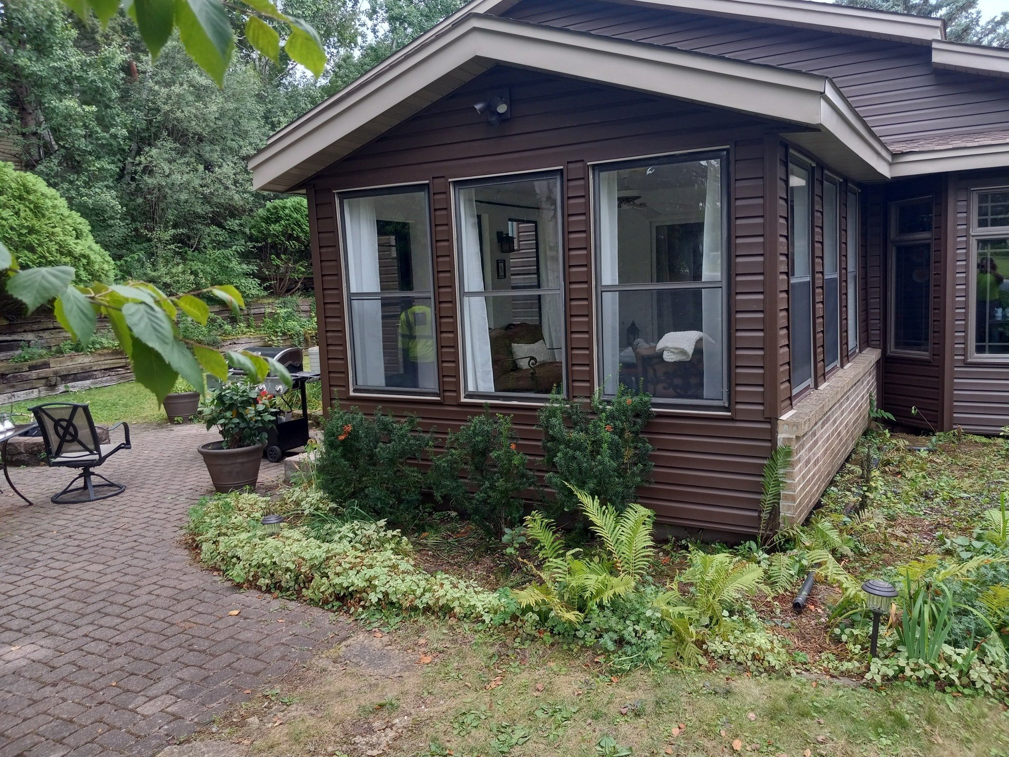 View of a house with large windows, brown siding, surrounded by lush green trees and plants in a backyard patio area with outdoor furniture.