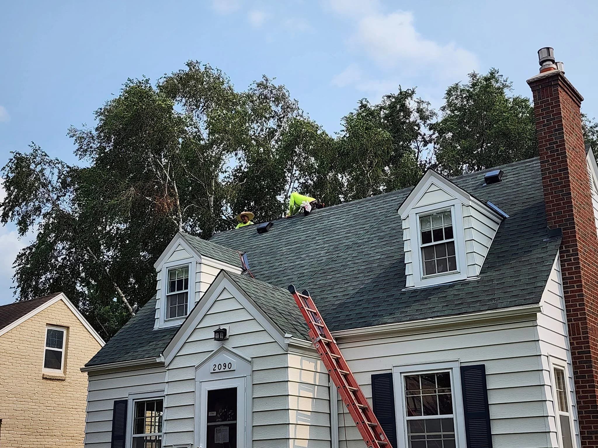 Two workers are on the roof of a white house, with one kneeling and working on the shingles and the other standing nearby. A red ladder leans against the house. The house has a brick chimney and several windows, and the scene is set against a clear sky with trees in the background.