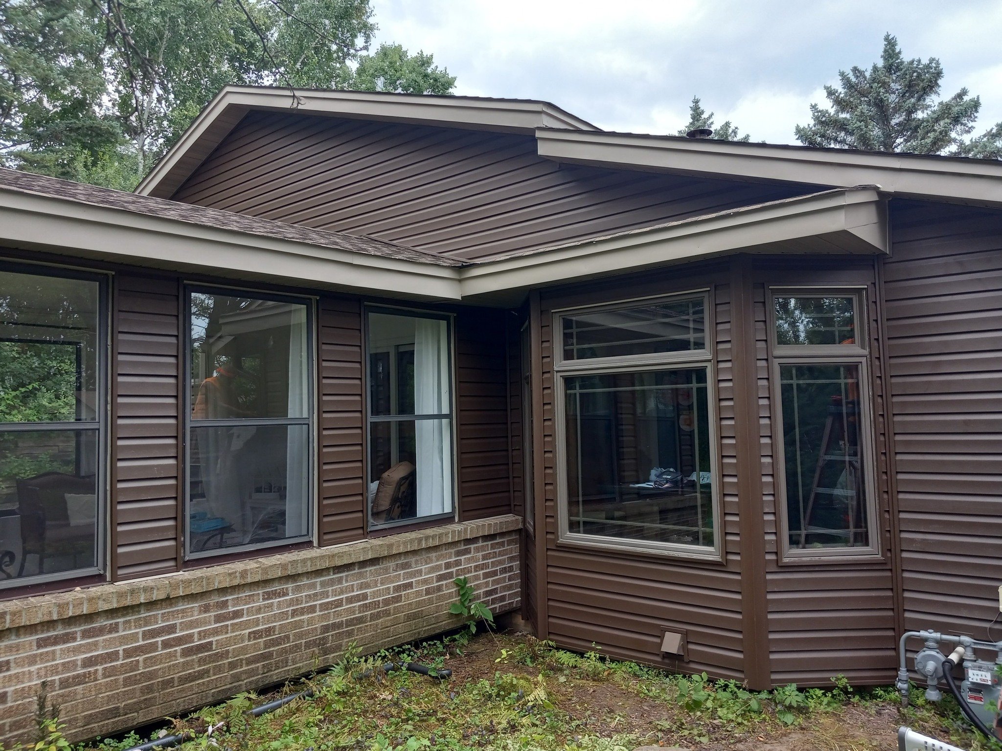 Back side of a house with large windows and brown siding. The house has a brick foundation and a small patch of garden in front with some green plants. Trees are visible in the background with cloudy skies.