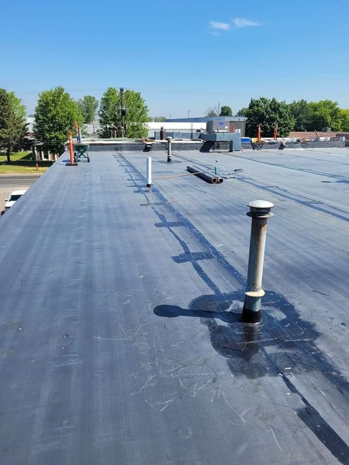 View of a flat commercial rooftop with HVAC vents, pipes, and flashing, under a clear blue sky with trees in the background.