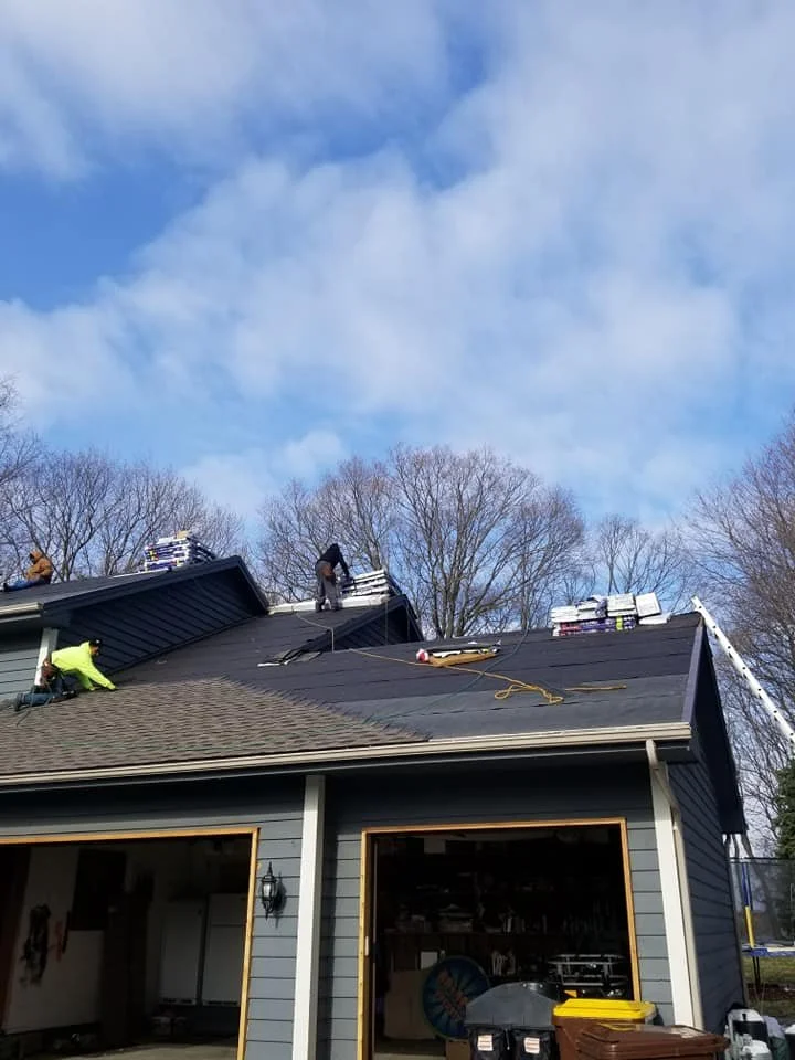 Construction workers installing roofing shingles on a house with a blue-gray exterior, during daytime with cloudy sky and leafless trees in the background.