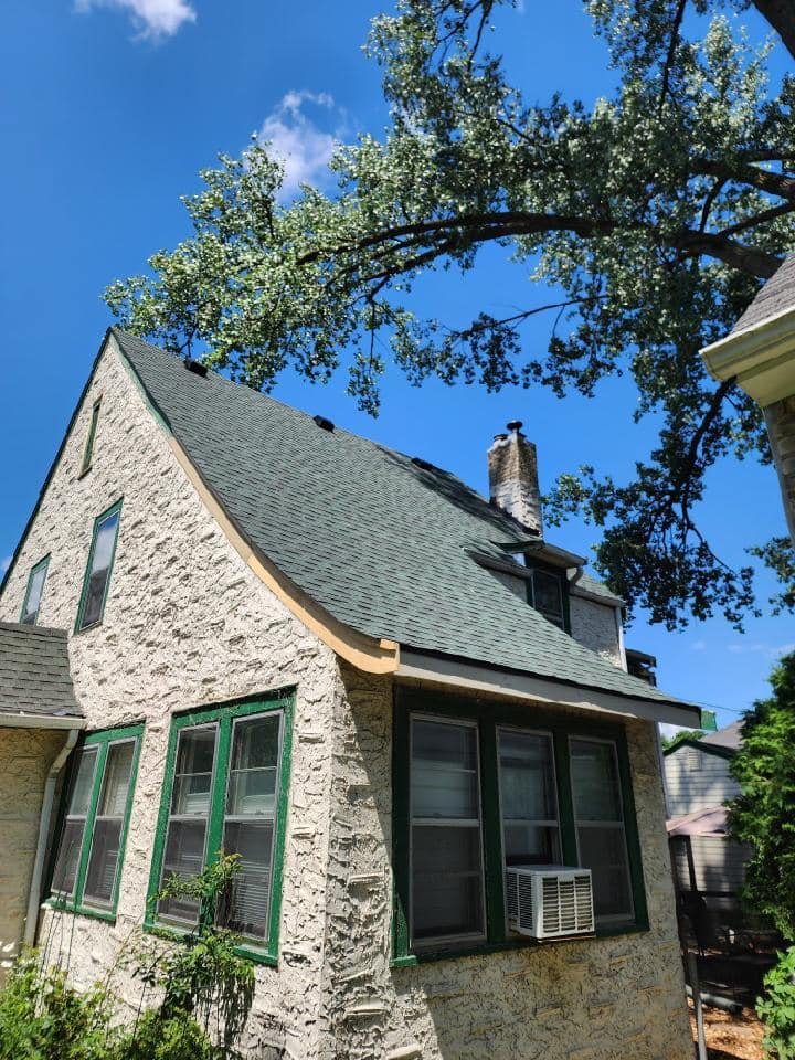 A house with a steep, gray shingled roof, cream-colored stucco walls, and green window frames. There is a tree with white blossoms and a blue sky with a few clouds in the background.