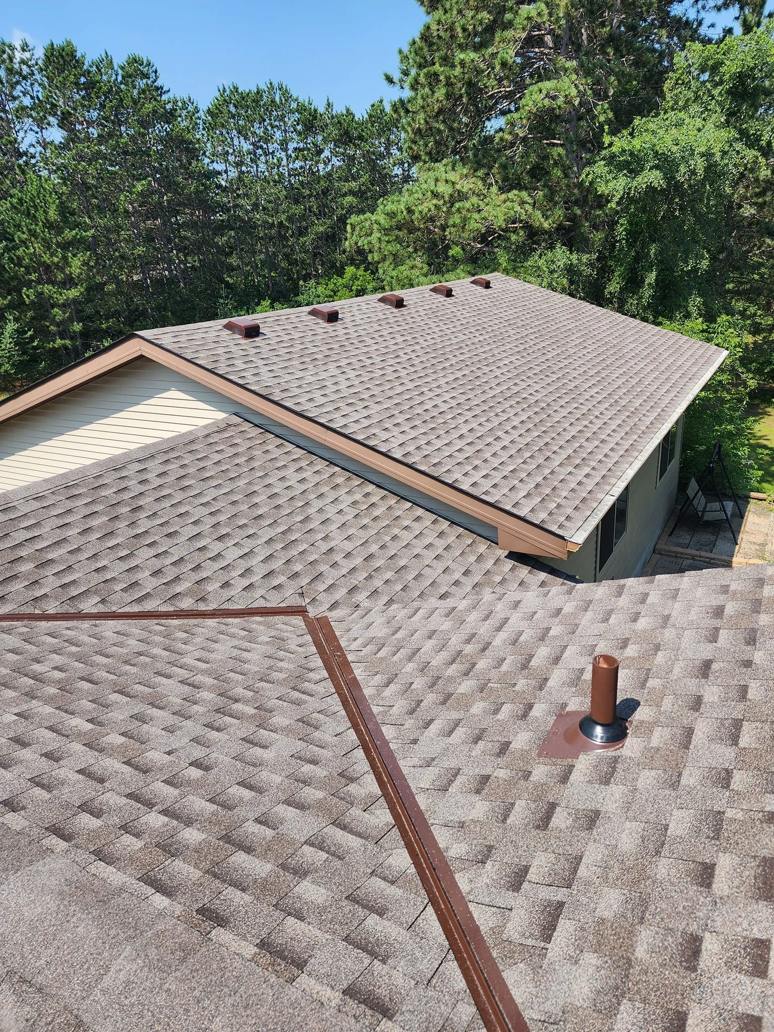 View of multiple house roofs covered with gray shingles, surrounded by tall green trees and a clear blue sky.