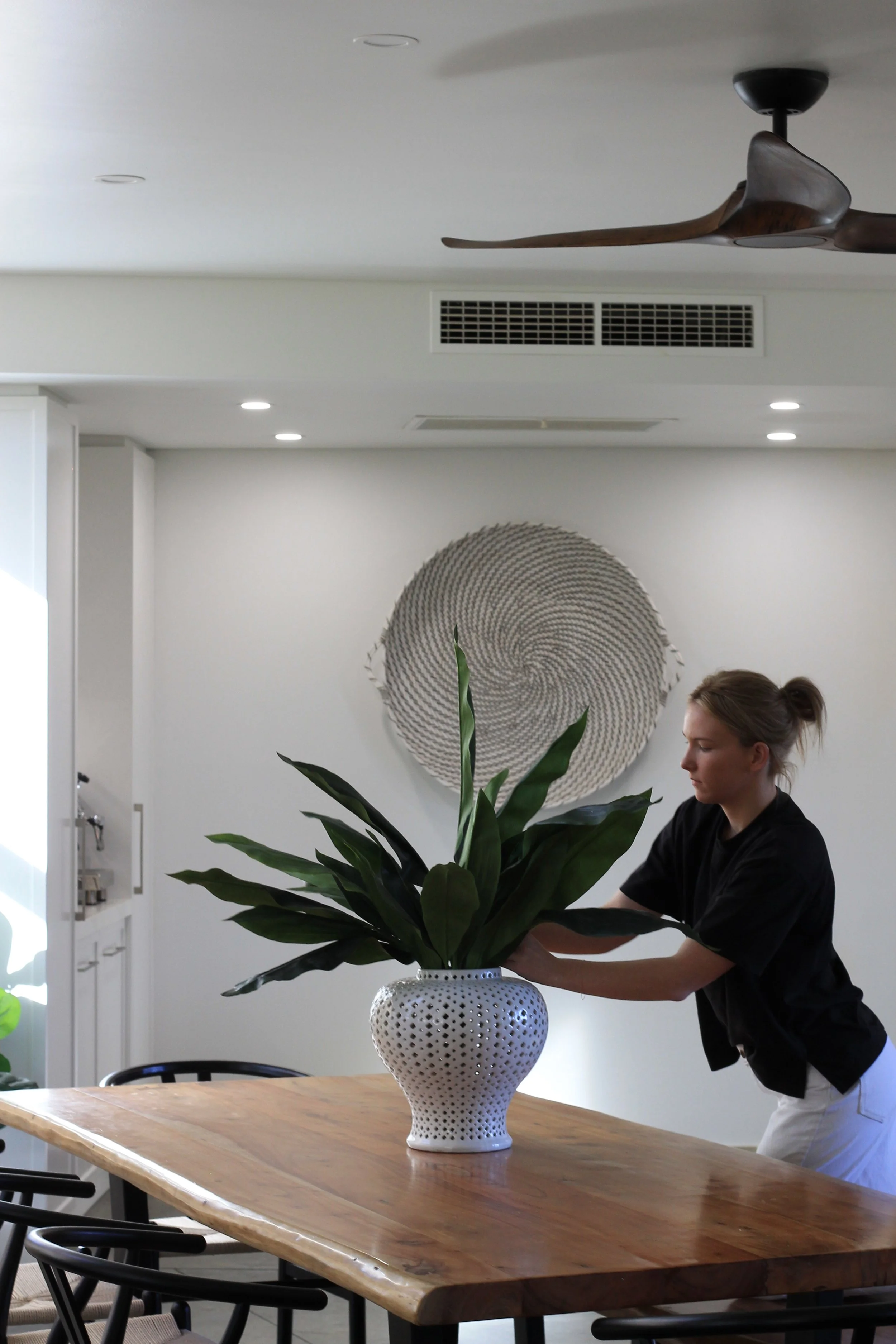 A woman is arranging a large potted green plant on a wooden dining table in a contemporary kitchen or dining area with white walls and a decorative round wall hanging.