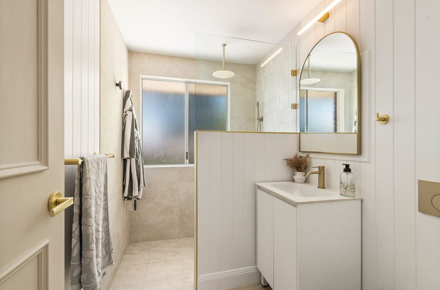 Modern bathroom with a glass-enclosed shower, a mirror with warm lighting, a white vanity with a gold faucet, and neutral decor including a soap dispenser, a small vase with dried flowers, and towels.