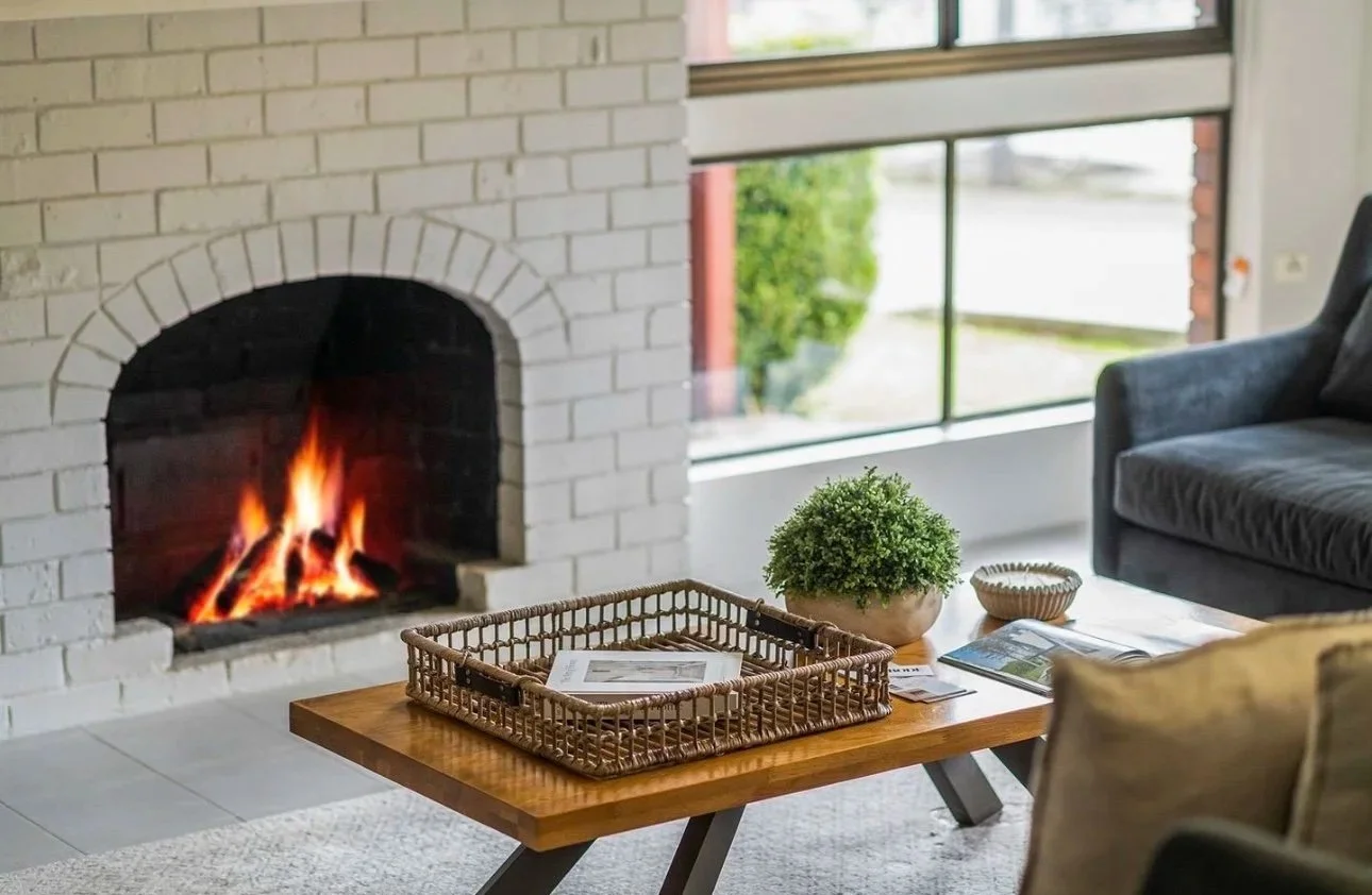 Living room with fireplace, lit with a warm fire, next to a window with a view of greenery outside, a coffee table with a plant, magazines, and a decorative bowl, and a gray couch.