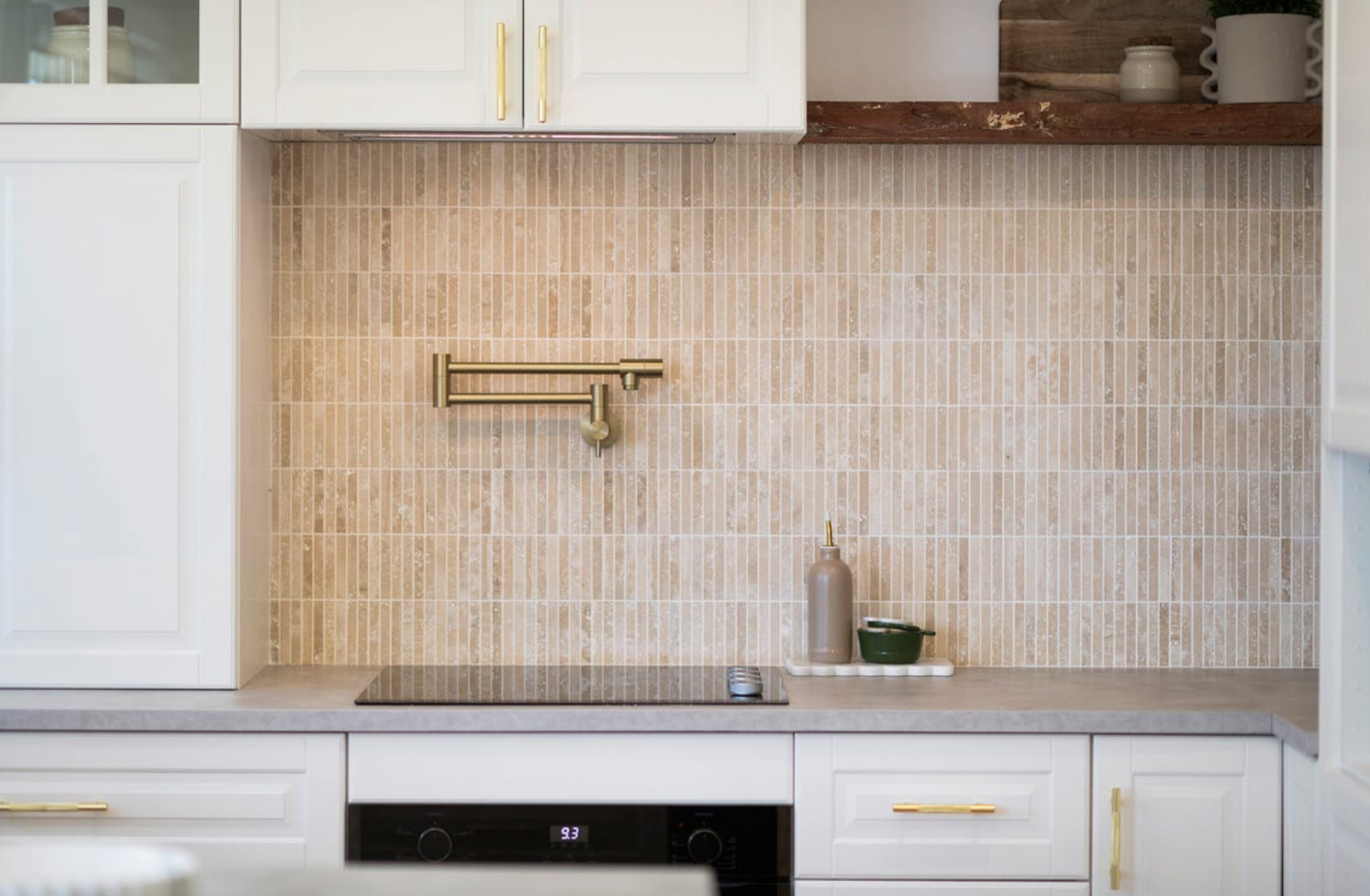 Modern kitchen with white cabinetry, gold handles, beige tiled backsplash, built-in oven, and a cooktop. Open shelves with white ceramics, a bottle, and small containers. Small countertop items include a green bowl and a bottle.