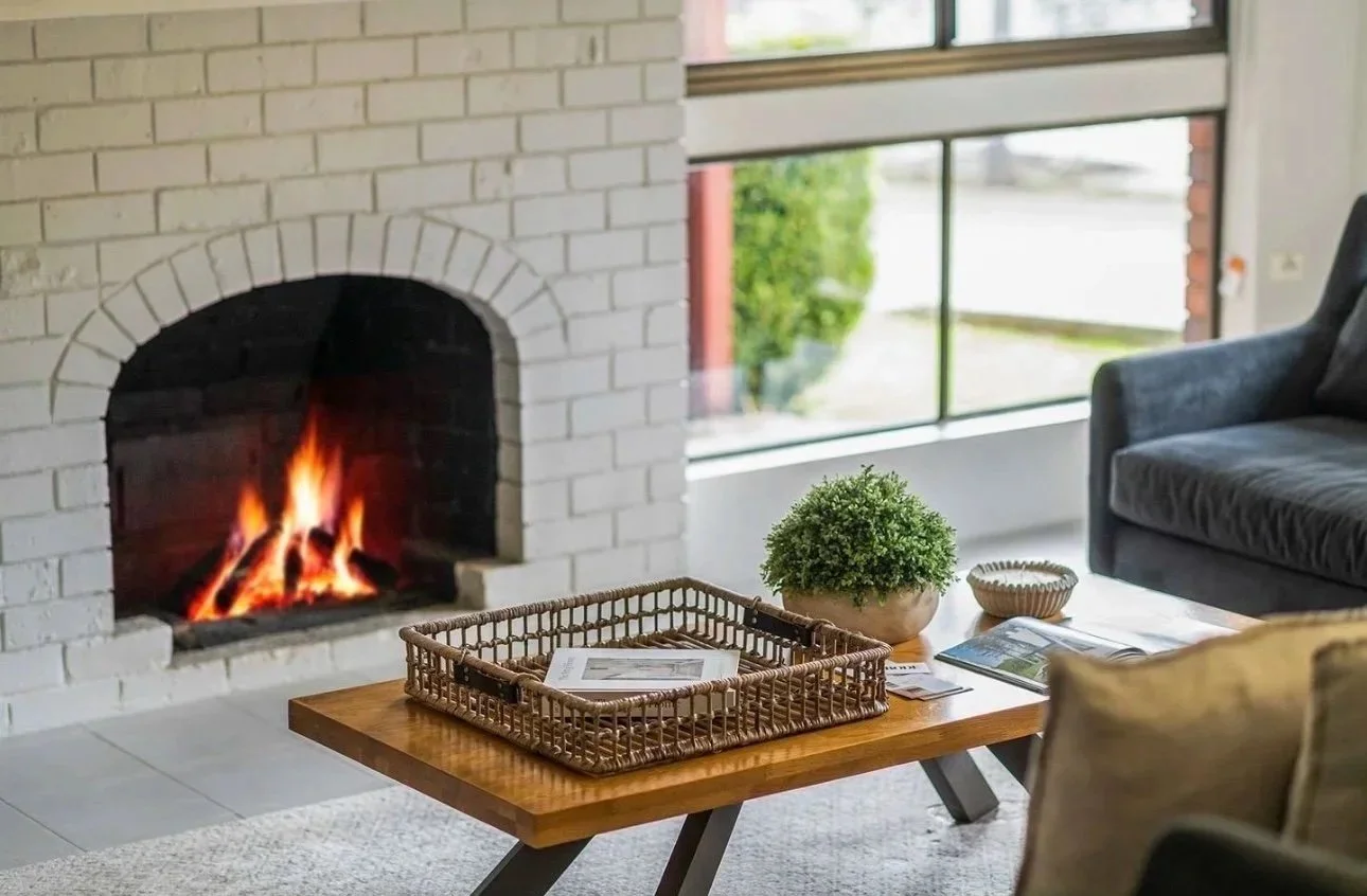 Living room with a lit fireplace, a wooden coffee table with a basket and magazines, a potted plant, a gray sofa, and large windows showing outdoor greenery.
