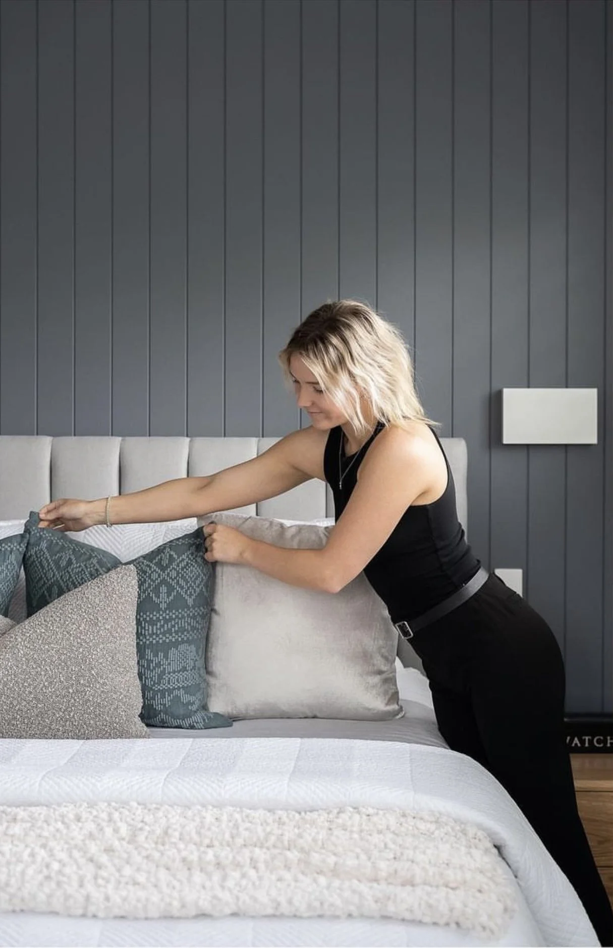 A woman arranging decorative pillows on a bed in a bedroom with a gray paneled wall.