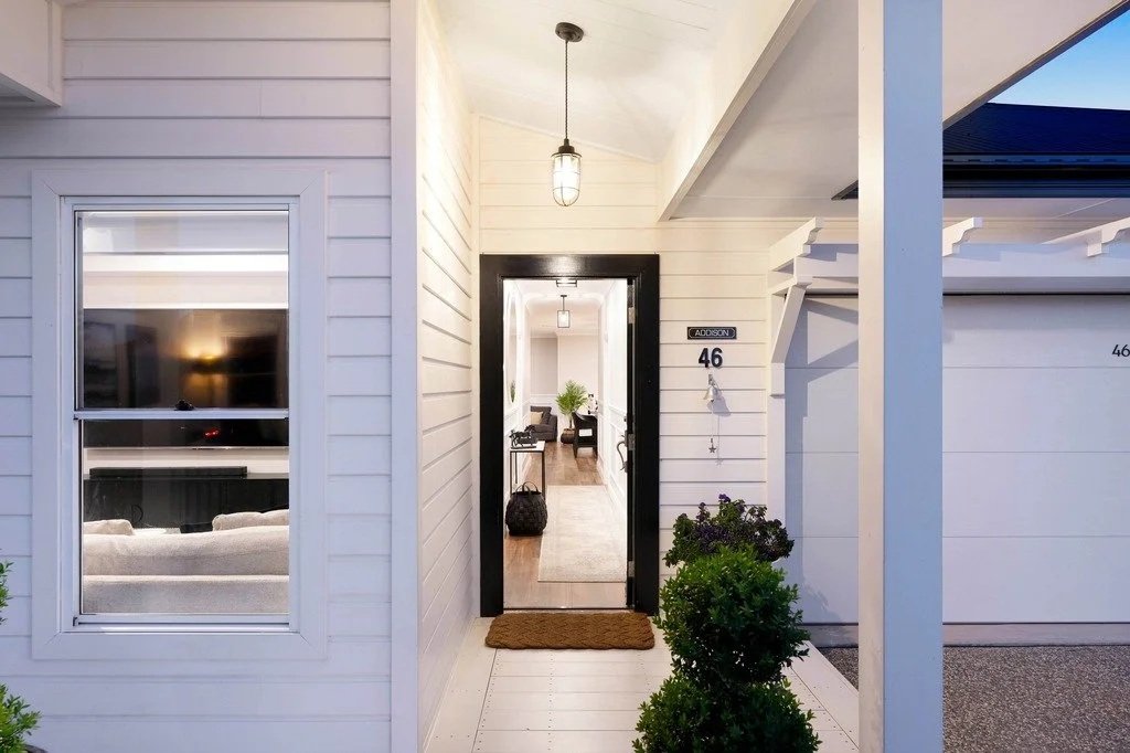 White house exterior with a black front door, house number 46, a porch with a brown doormat, and some greenery. Inside, a glimpse of a living room with a TV, beige sofa, and a room with beige walls and black furniture.