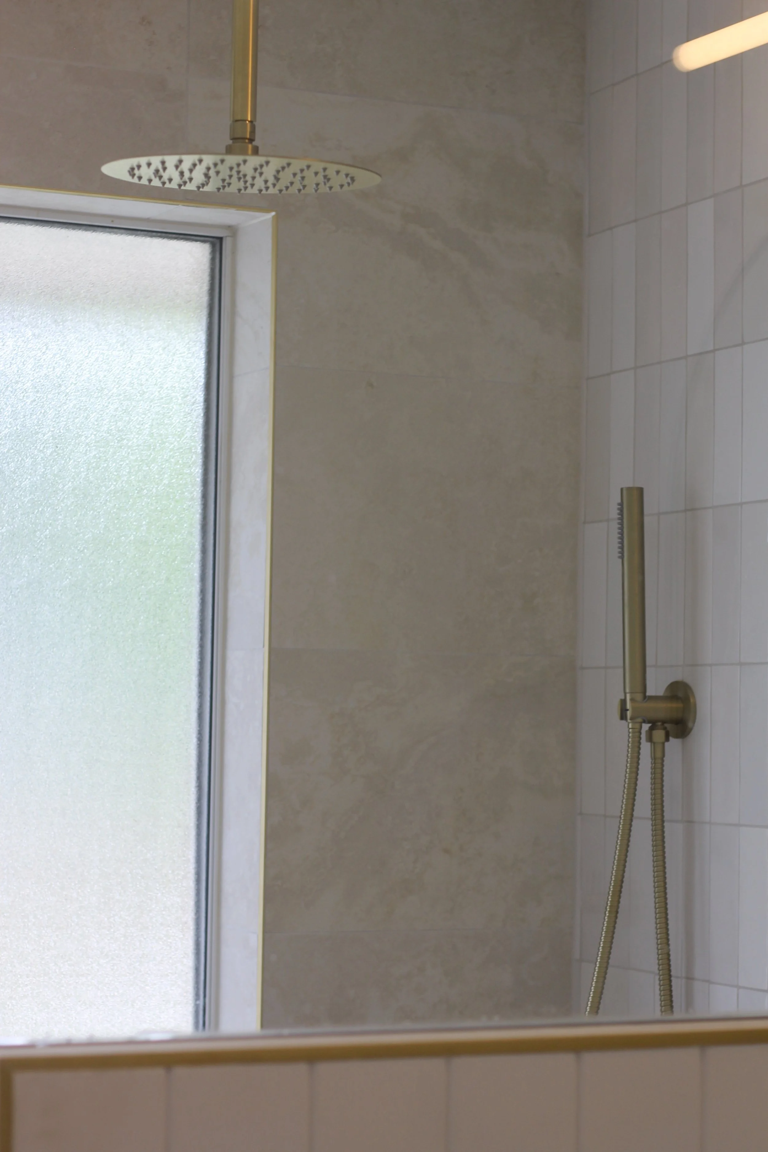 A shower with a rain shower head attached to the ceiling, a soap holder, and a handheld shower hose, with frosted glass window on the left and tiled wall on the right.