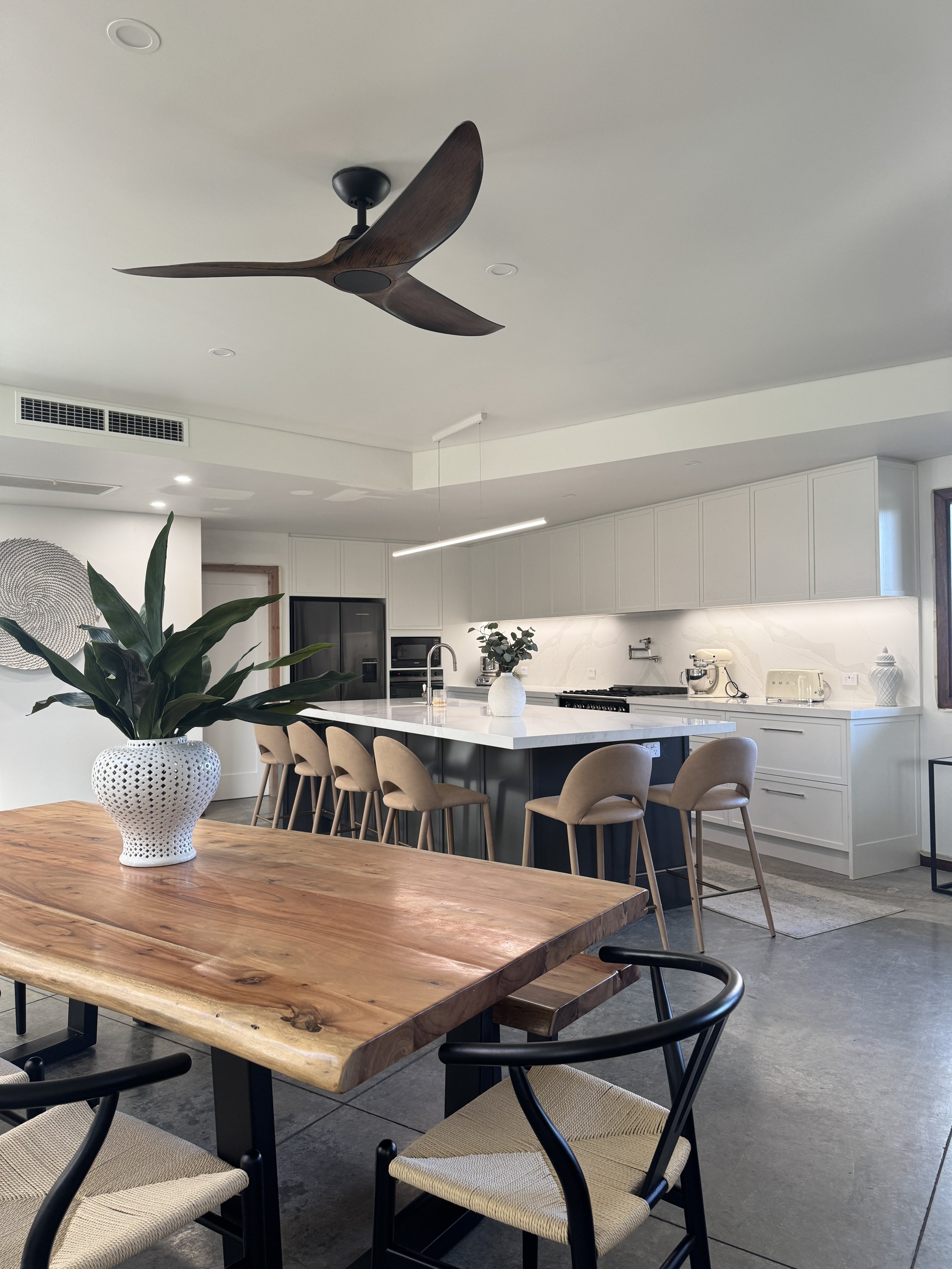 Modern kitchen and dining area with a wooden table, beige chairs, a white kitchen island with a vase of green plants, white cabinets, and a ceiling fan.