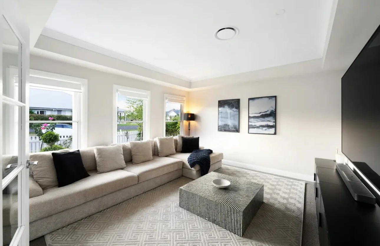 Bright living room with large beige sectional sofa, black pillows, a striped coffee table, a patterned area rug, four windows, artwork on the wall, a floor lamp, and a large flat-screen TV.
