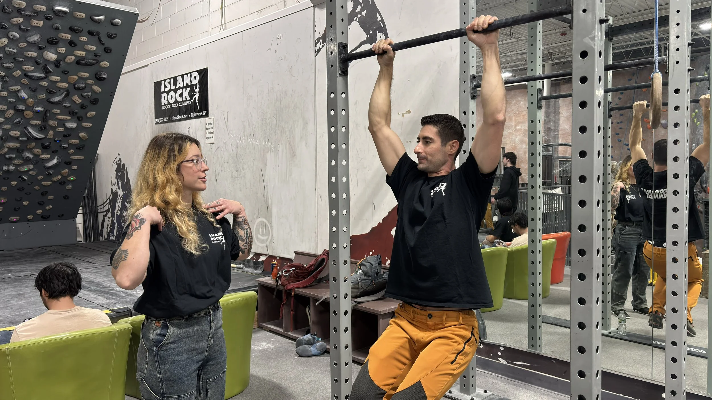 A man is doing pull-ups on a bar in an indoor rock climbing gym. A woman is standing nearby, talking to him. The gym has a large climbing wall and other people in the background, some sitting and some climbing.