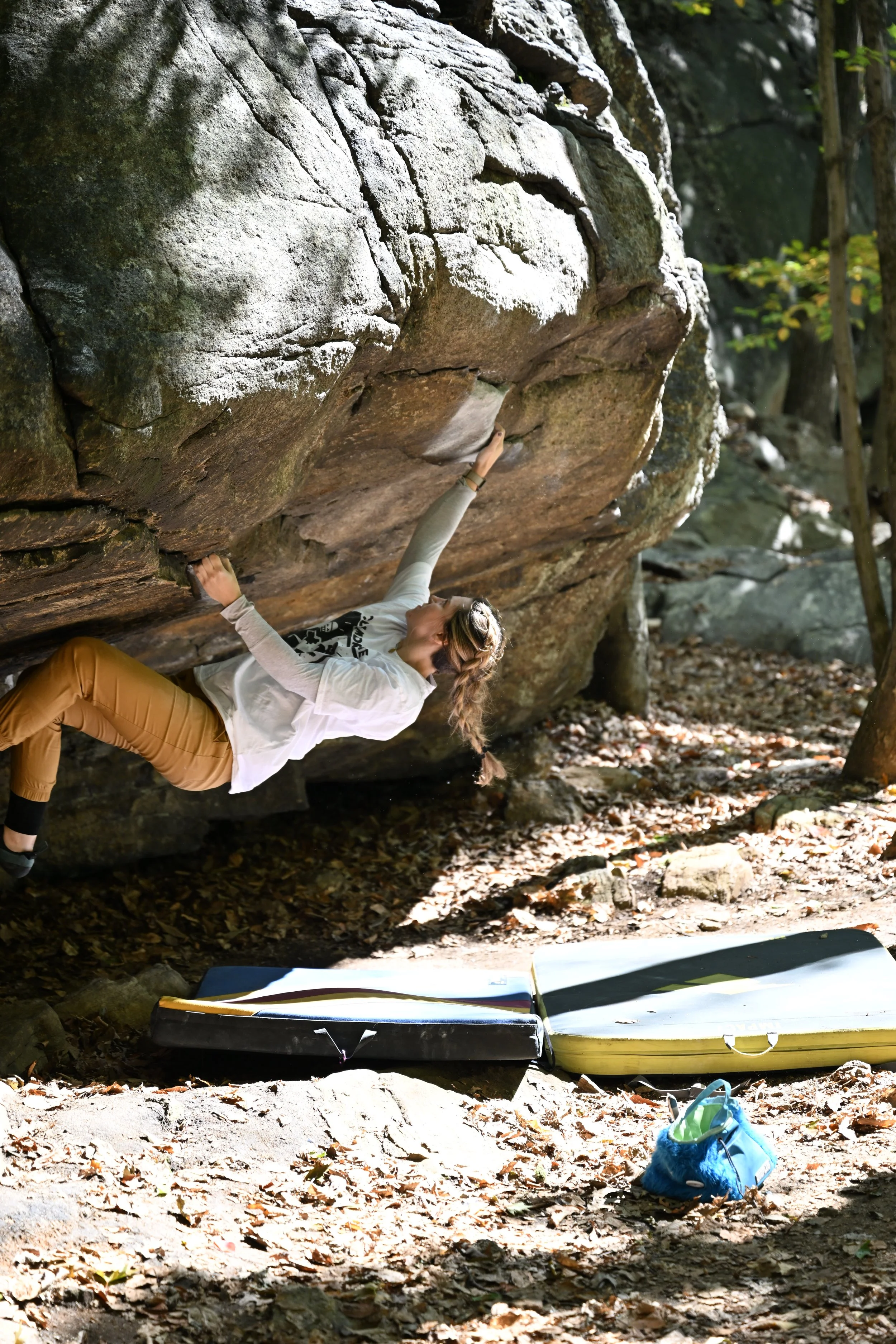 A woman bouldering outdoors under a large rock formation, with climbing shoes, crash pads, and hiking gear nearby, surrounded by fall leaves and trees.