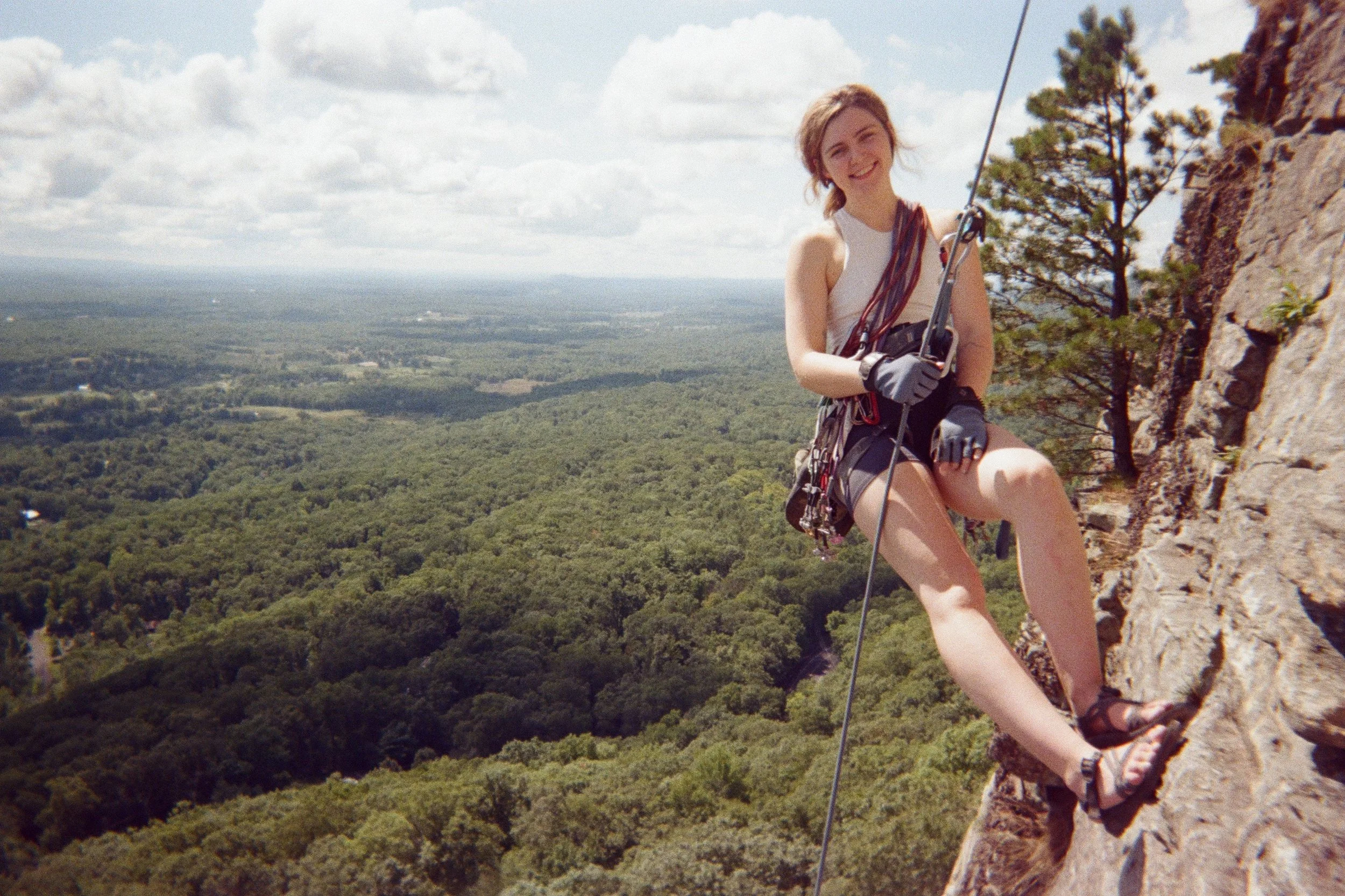 Woman climbing a rock face on a hiking trail with a scenic view of green forests and cloudy sky in the background.
