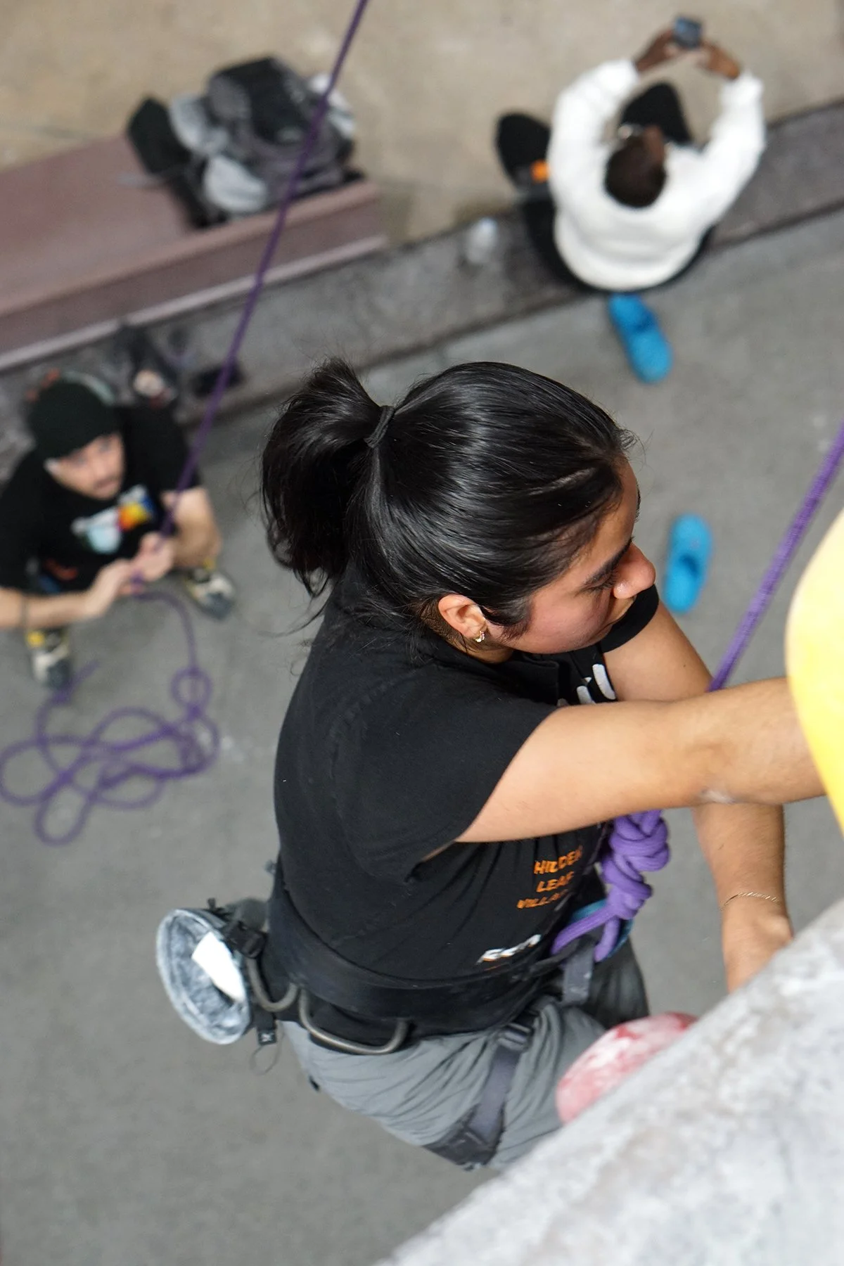 A woman climbing a rock wall at Island Rock Climbing Gym, viewed from above, with other climbers and equipment in the background.