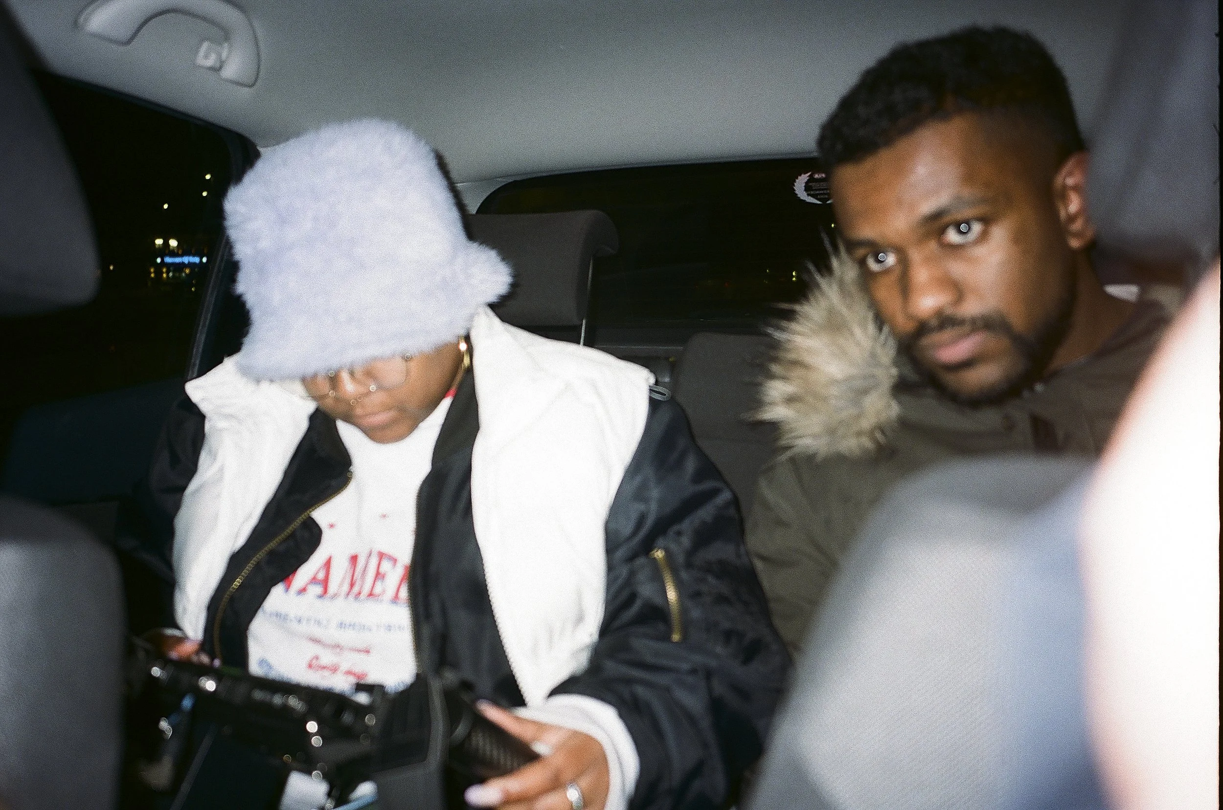 Two people sitting in the backseat of a car at night, one wearing a gray beanie and glasses, looking down at a camera, the other looking at the camera with a fur-lined hood