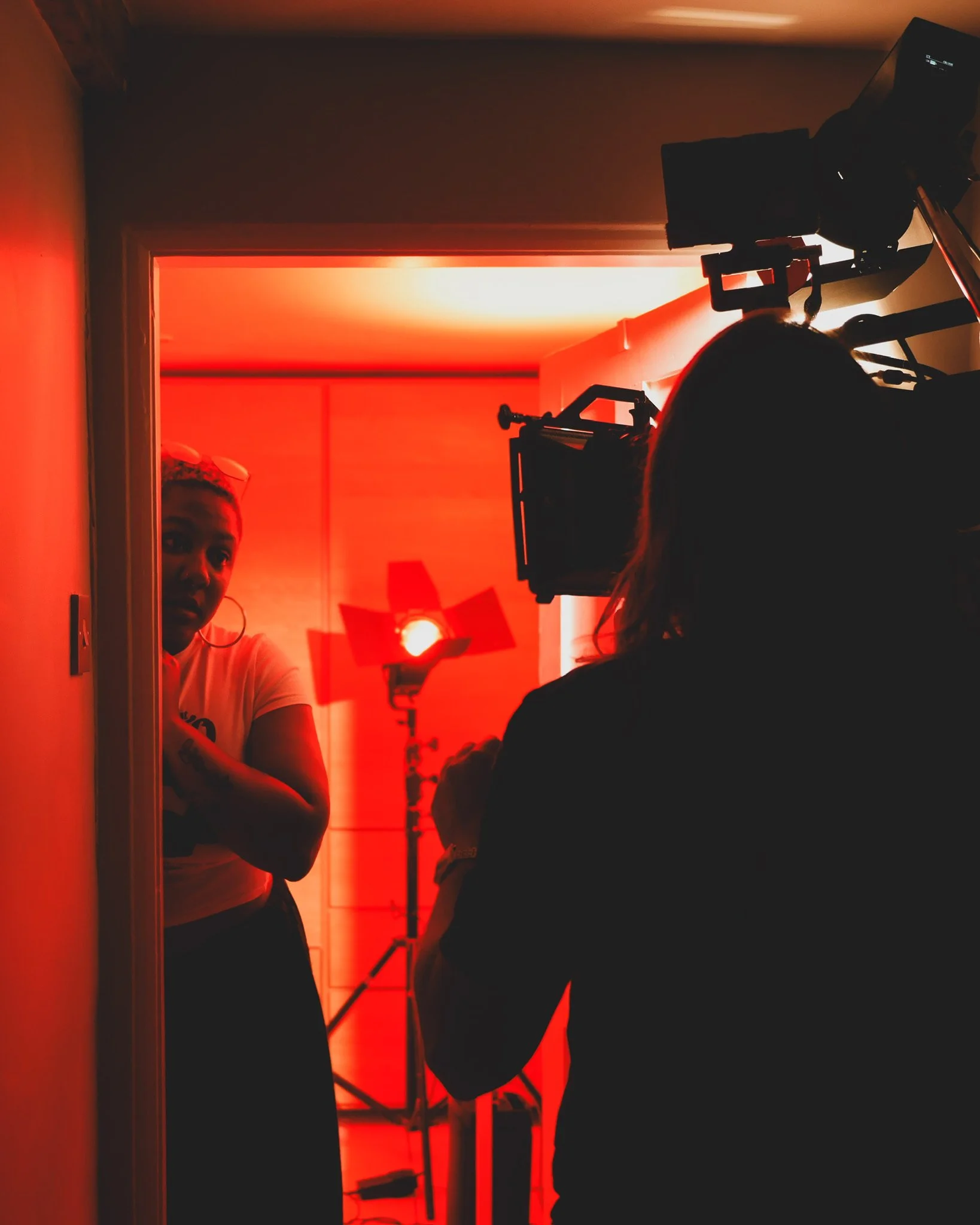 A woman being filmed by a person with a camera, with red lighting and studio equipment in the background.