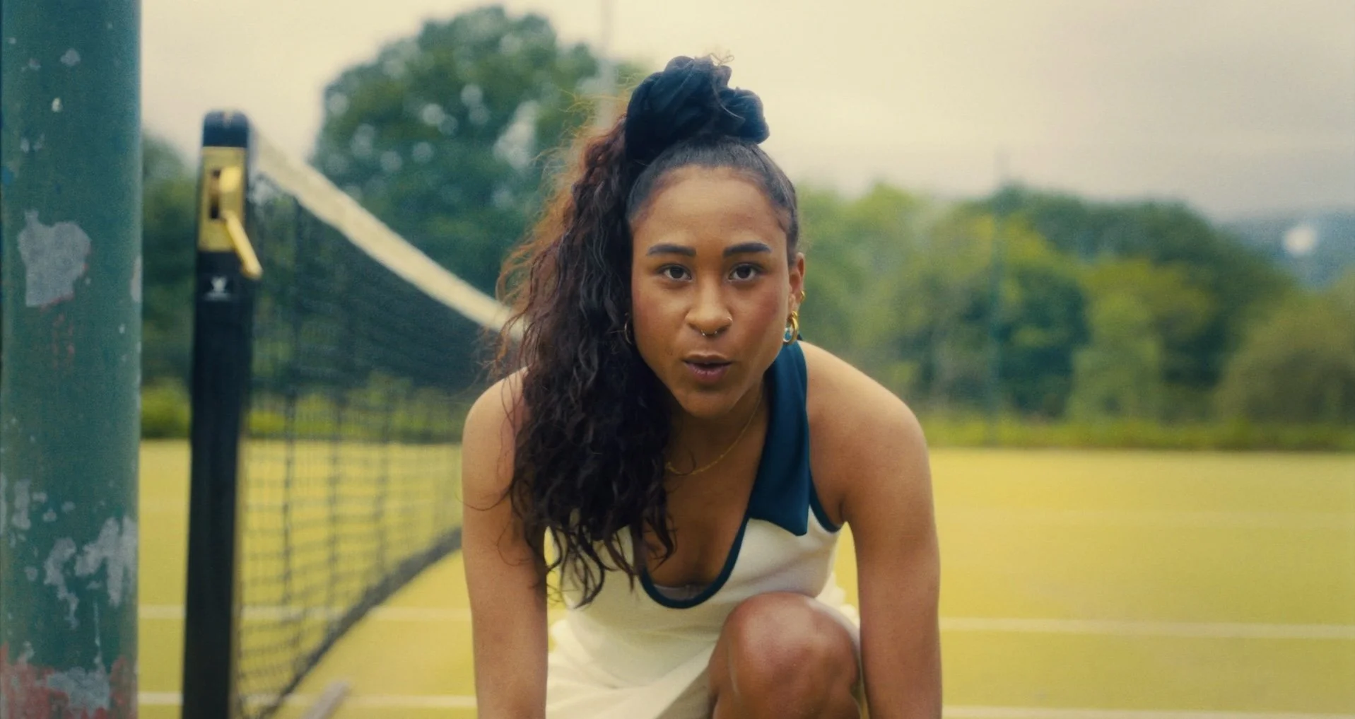 A woman with long curly hair in a high ponytail, wearing a white sports dress with teal accents, crouching on a tennis court.