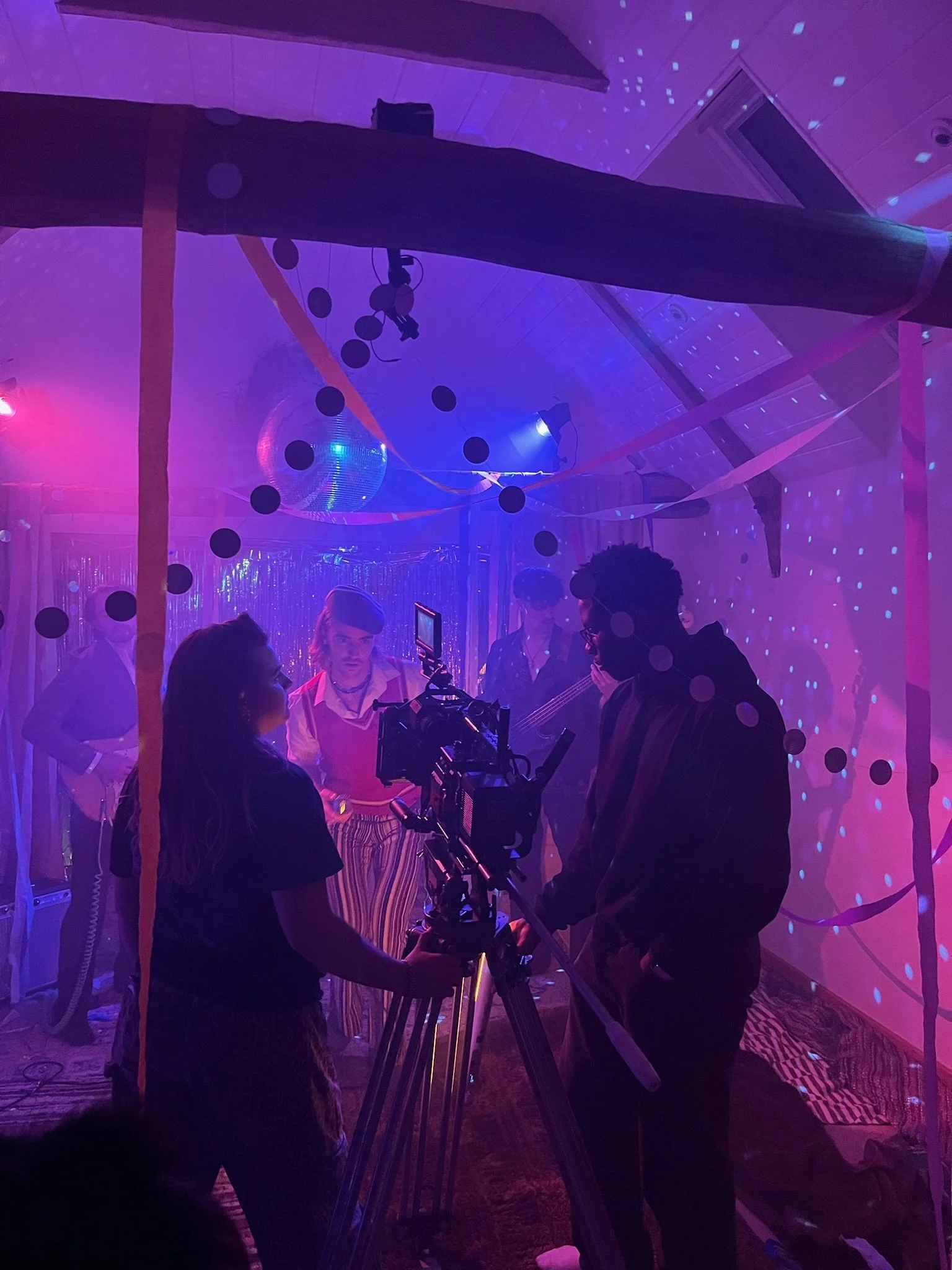A film crew filming a scene with a group of people inside a decorated room with purple and pink lighting, streamers, balloons, and a disco ball.