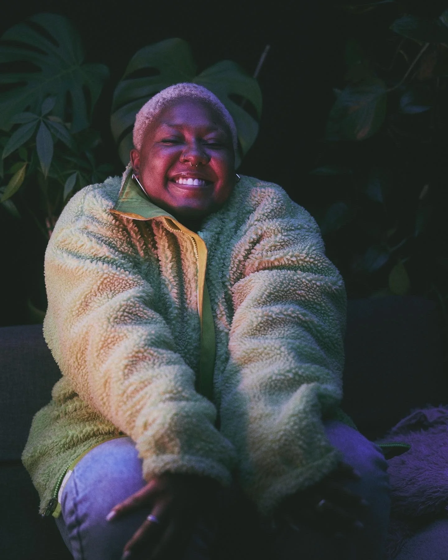A person with short blond hair smiling widely while sitting on a couch, surrounded by dark green plants.