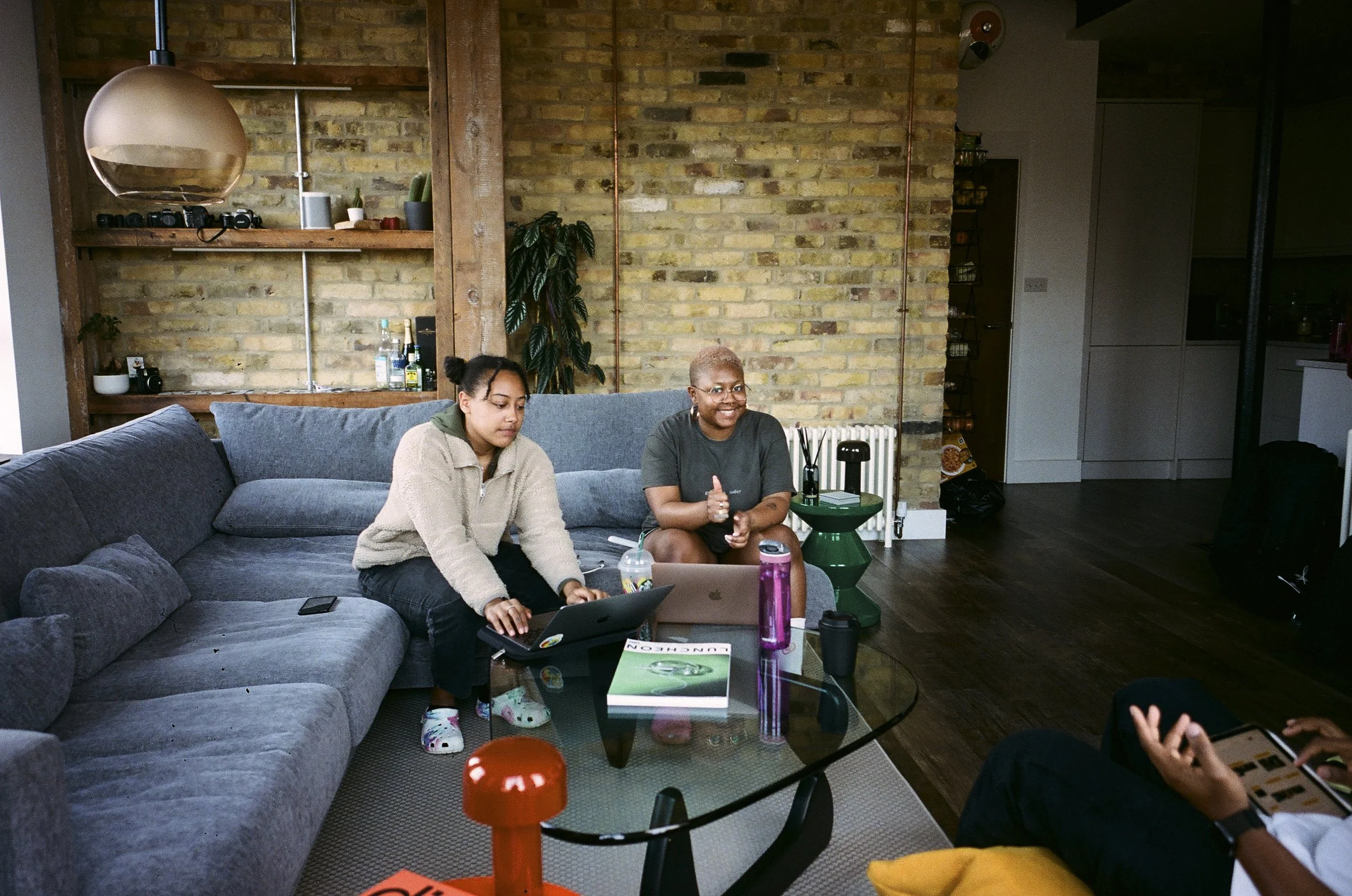 Two women sitting on a gray sofa in a modern living room with brick walls, one woman working on a laptop and the other smiling and clapping, with a glass coffee table in front of them and another person partially visible on the right side of the imag