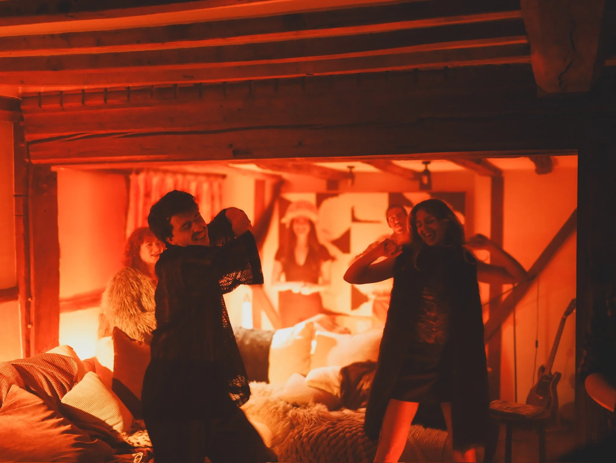 Four women dancing and enjoying themselves in a cozy, warmly lit room with wooden beams, pillows, a guitar, and wall art.
