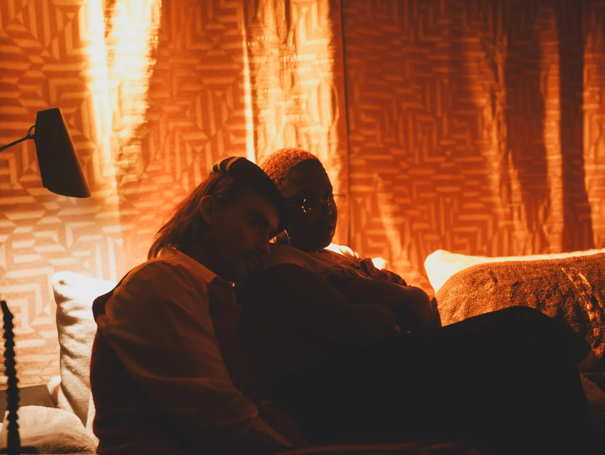 Two people sitting on a bed in a dimly lit room with orange patterned curtains behind them. One person has light hair and is wearing a shirt, while the other has short hair, glasses, and darker skin.