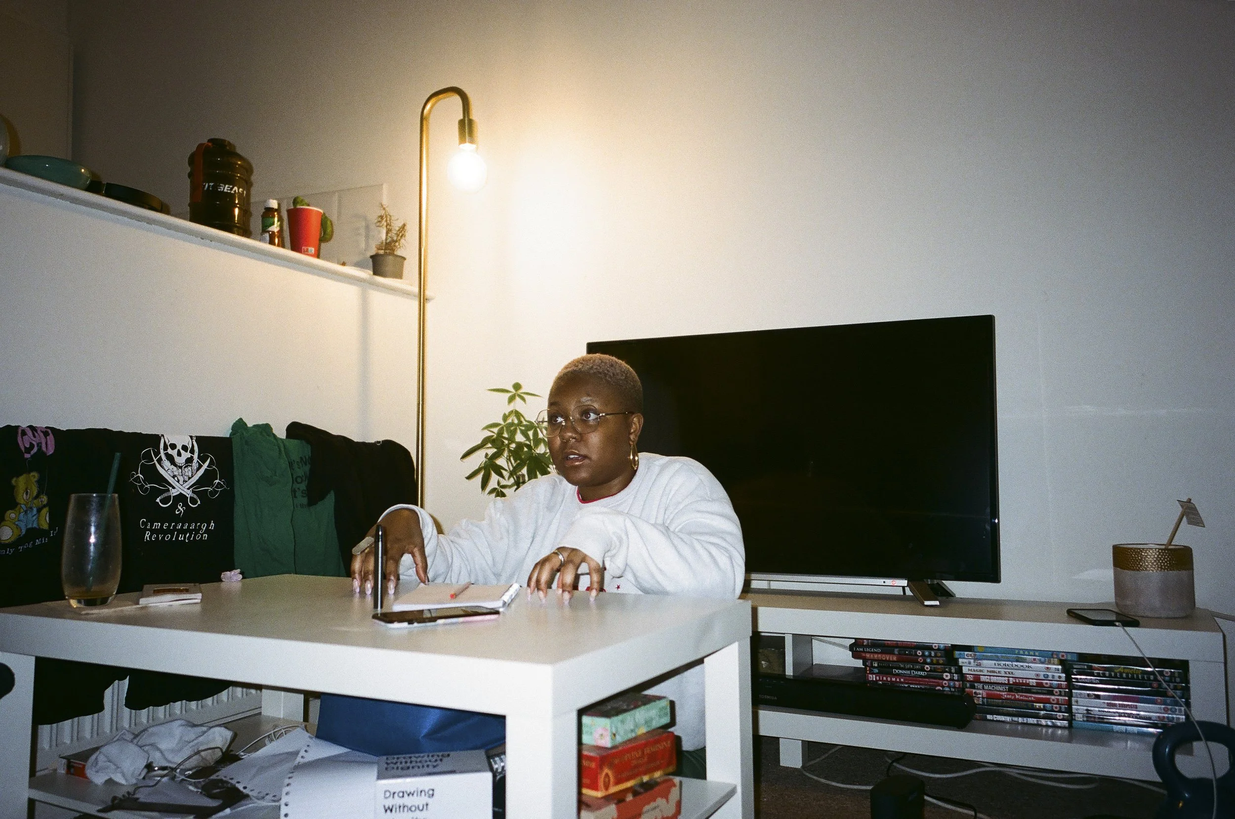 A woman with glasses sitting at a white table in a living room with a TV and bookshelf, wearing a white sweatshirt, with notebooks and a glass of drink in front of her.