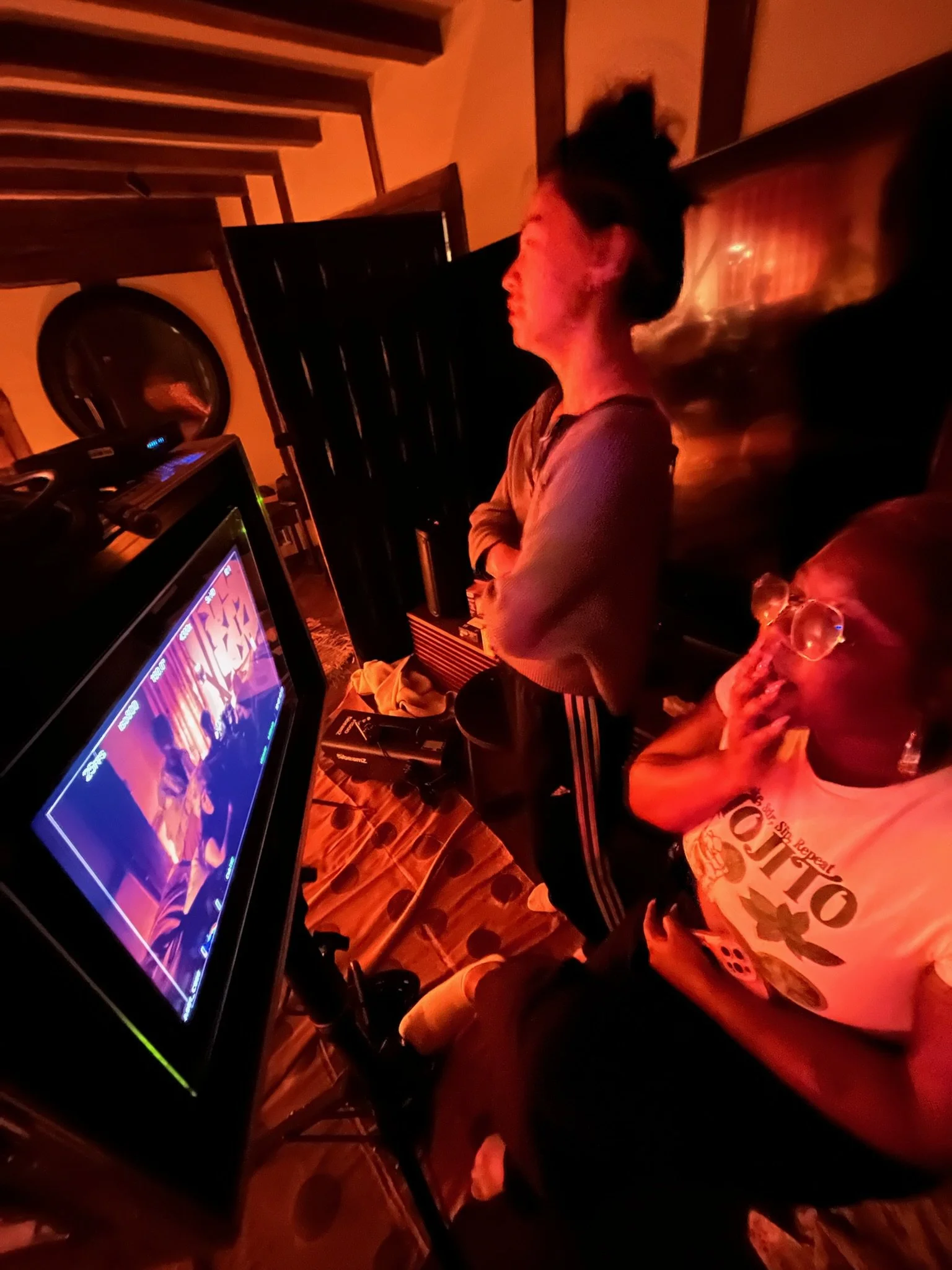 Two women watching a karaoke screen in a dimly lit room with warm orange and red lighting.