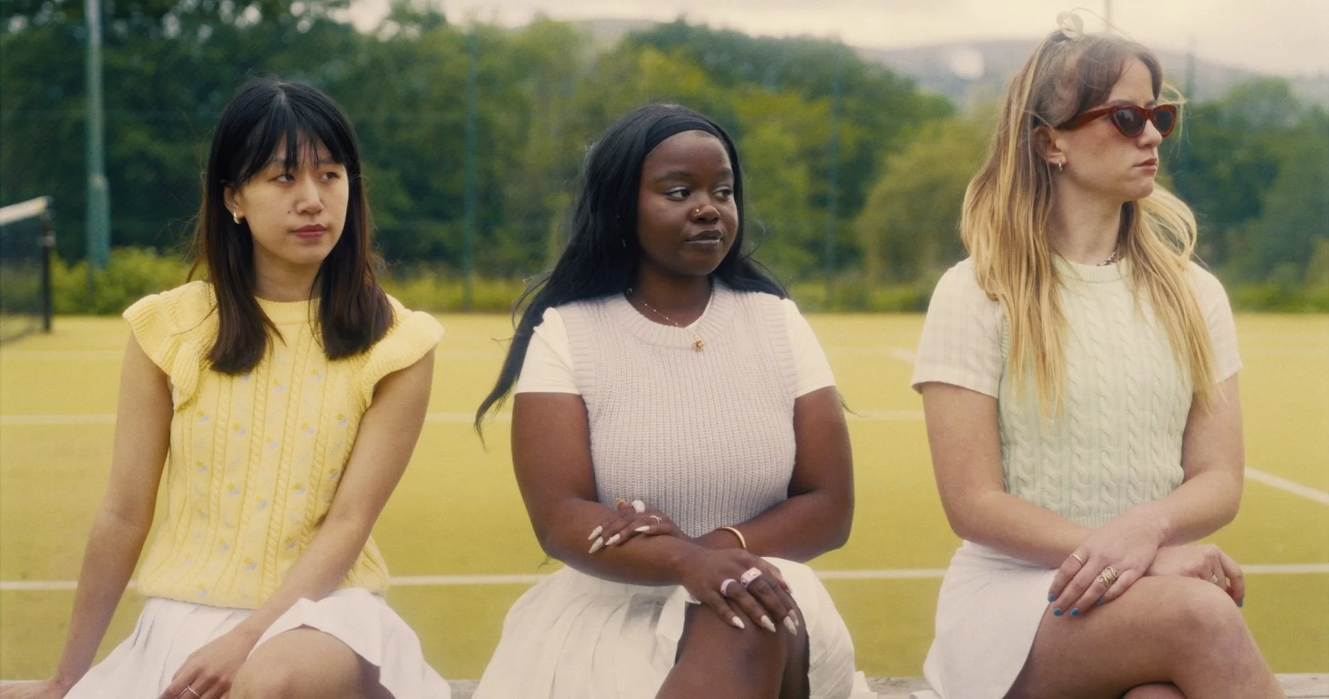 Three women sitting outdoors on a yellow tennis court with green trees in the background.