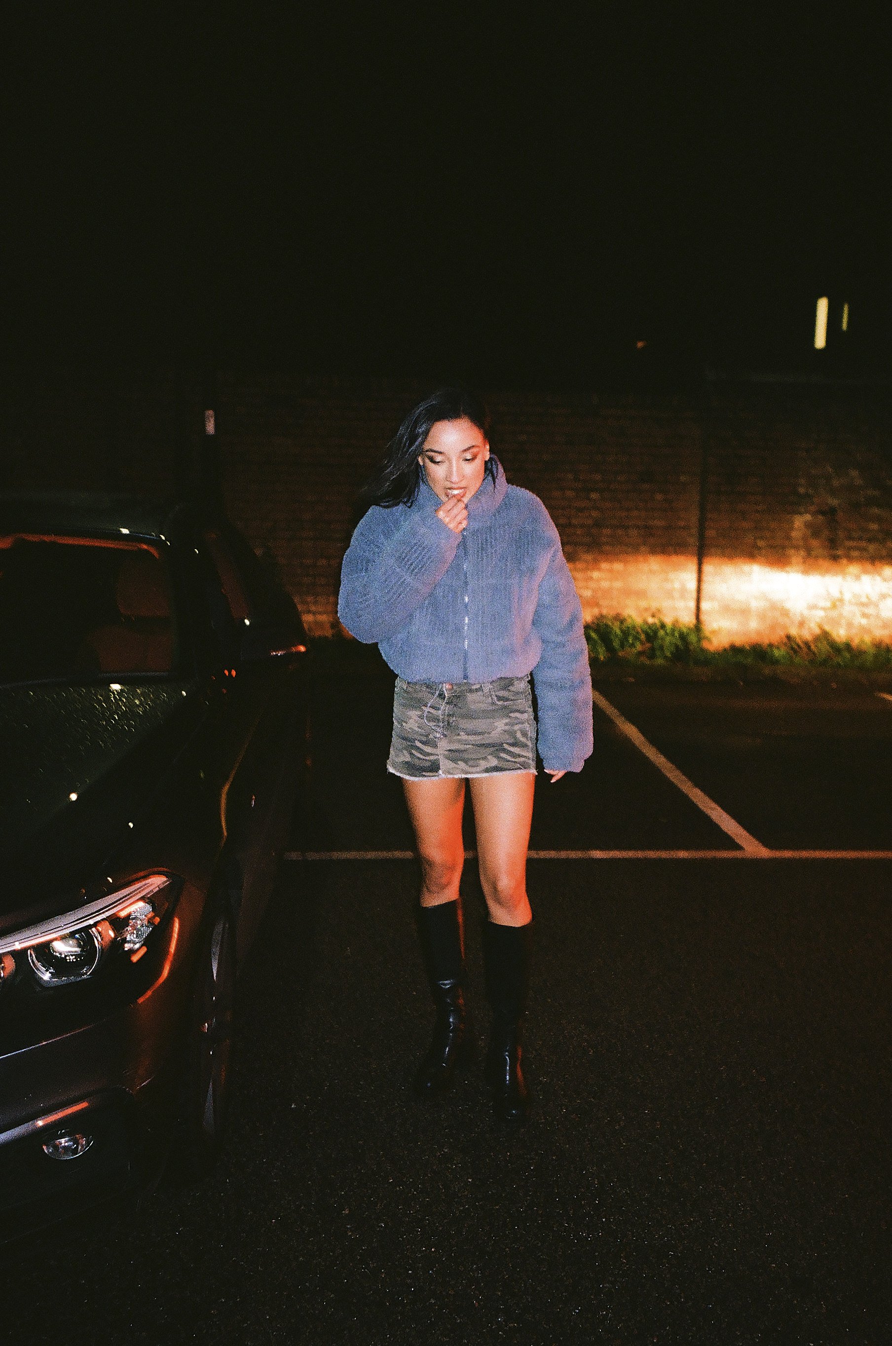A woman stands outdoors at night near a car, wearing a blue jacket, a camouflage skirt, and black knee-high boots, with a brick wall in the background.