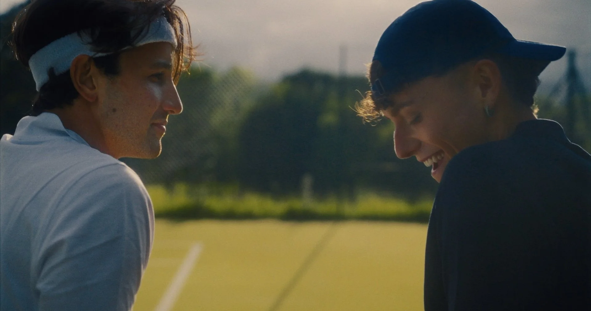 Two young men with curly hair smiling and talking on a tennis court, one wearing a white shirt and headband, the other wearing a dark shirt and cap.