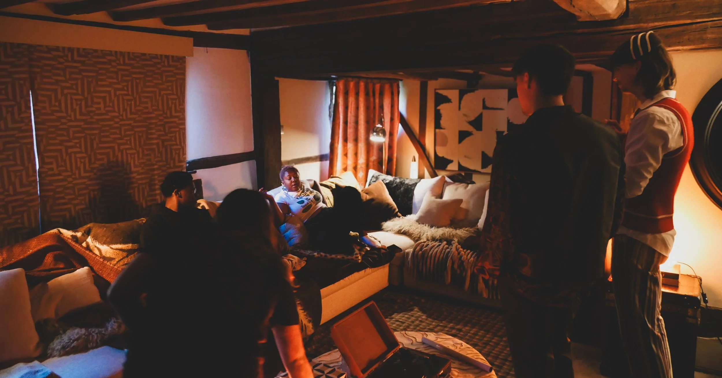 A group of people gathered in a cozy, dimly-lit living room with exposed wooden beams, colorful curtains, and abstract wall art. Some are sitting on a sofa and others are standing, engaging in conversation.