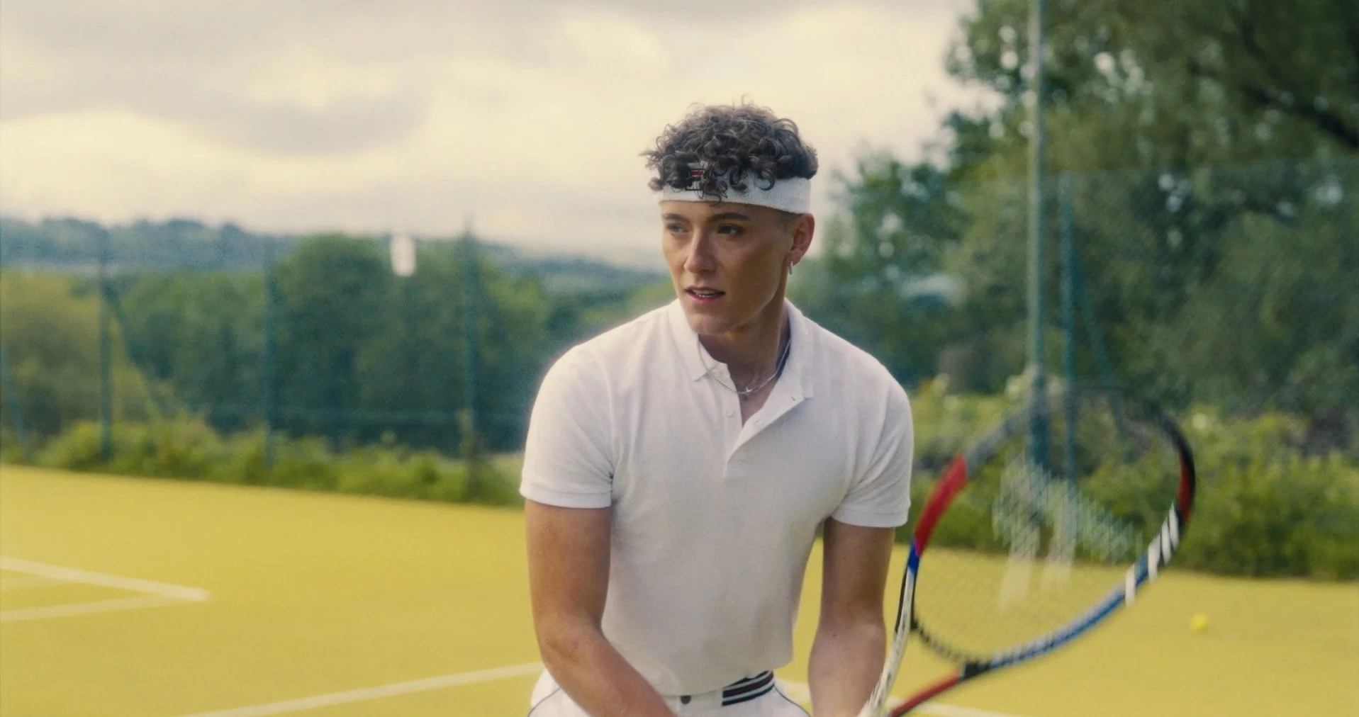 A woman with curly hair wearing a white headband and white athletic shirt, holding a tennis racket on a tennis court.