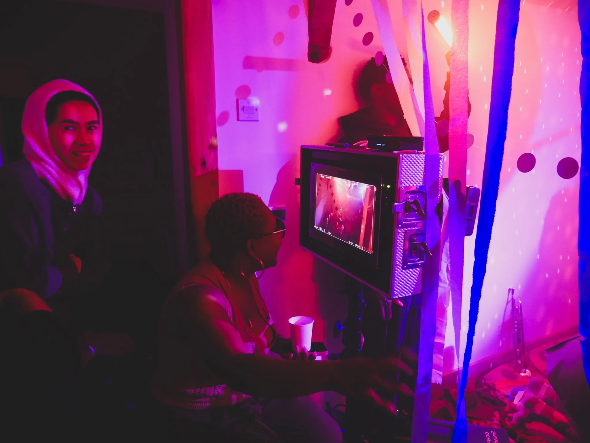 Three people in a dark room with pink and purple lights, two women and one man, with one woman sitting at a computer monitor, the other standing and smiling, in a setting with decorations and a curtain separating two areas.