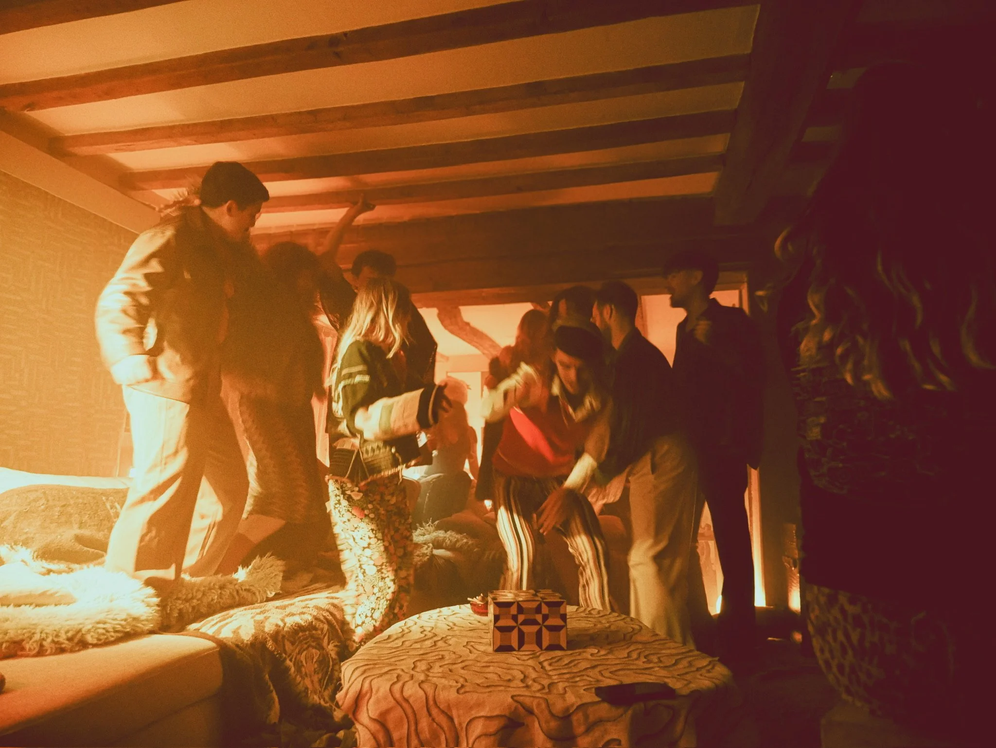 Group of people dancing and socializing in a dimly lit room with wooden beams on the ceiling, a patterned coffee table, and a plush sofa.