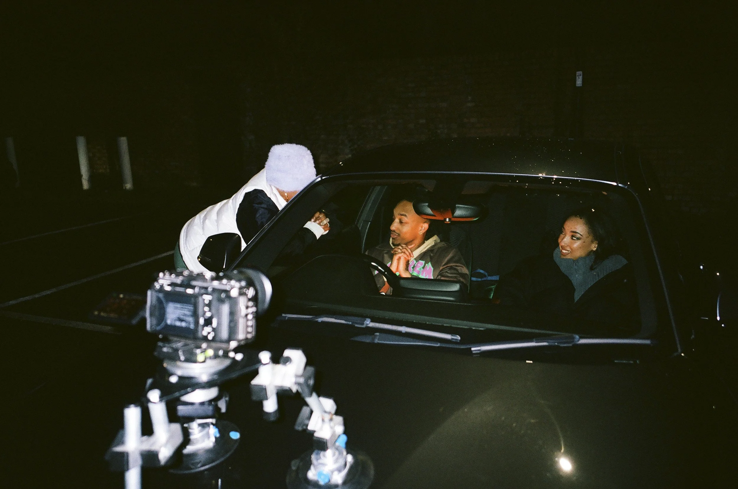 A person standing outside a car talking to two people sitting inside at night, with camera equipment on a tripod in the foreground.