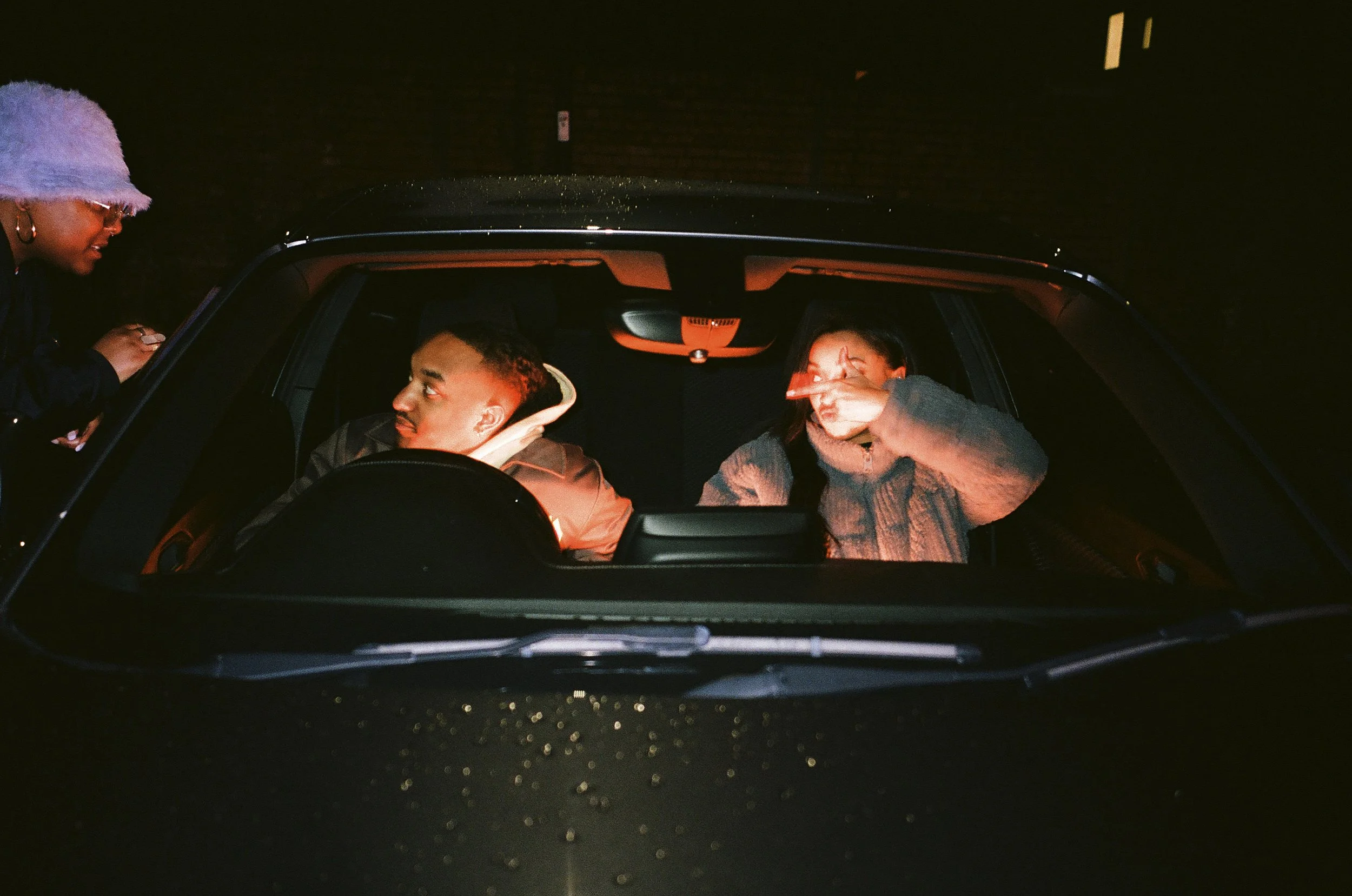 Two women and a man inside a car at night, with one woman outside the car leaning in to talk.