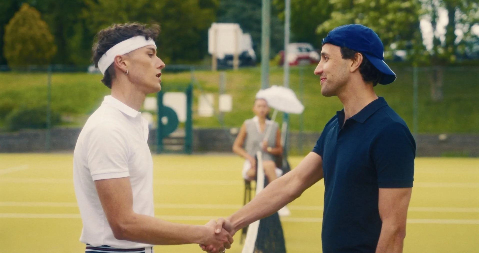 A man and a woman shake hands on a tennis court with green trees in the background. The woman is wearing a white tennis outfit with a headband, and the man is dressed in a navy shirt and a backwards cap.