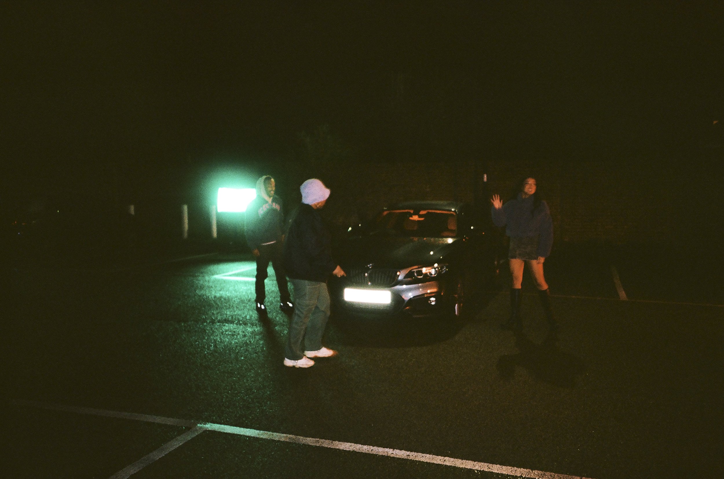 Three people standing around a black car in a dark parking lot at night, with a green neon light in the background.