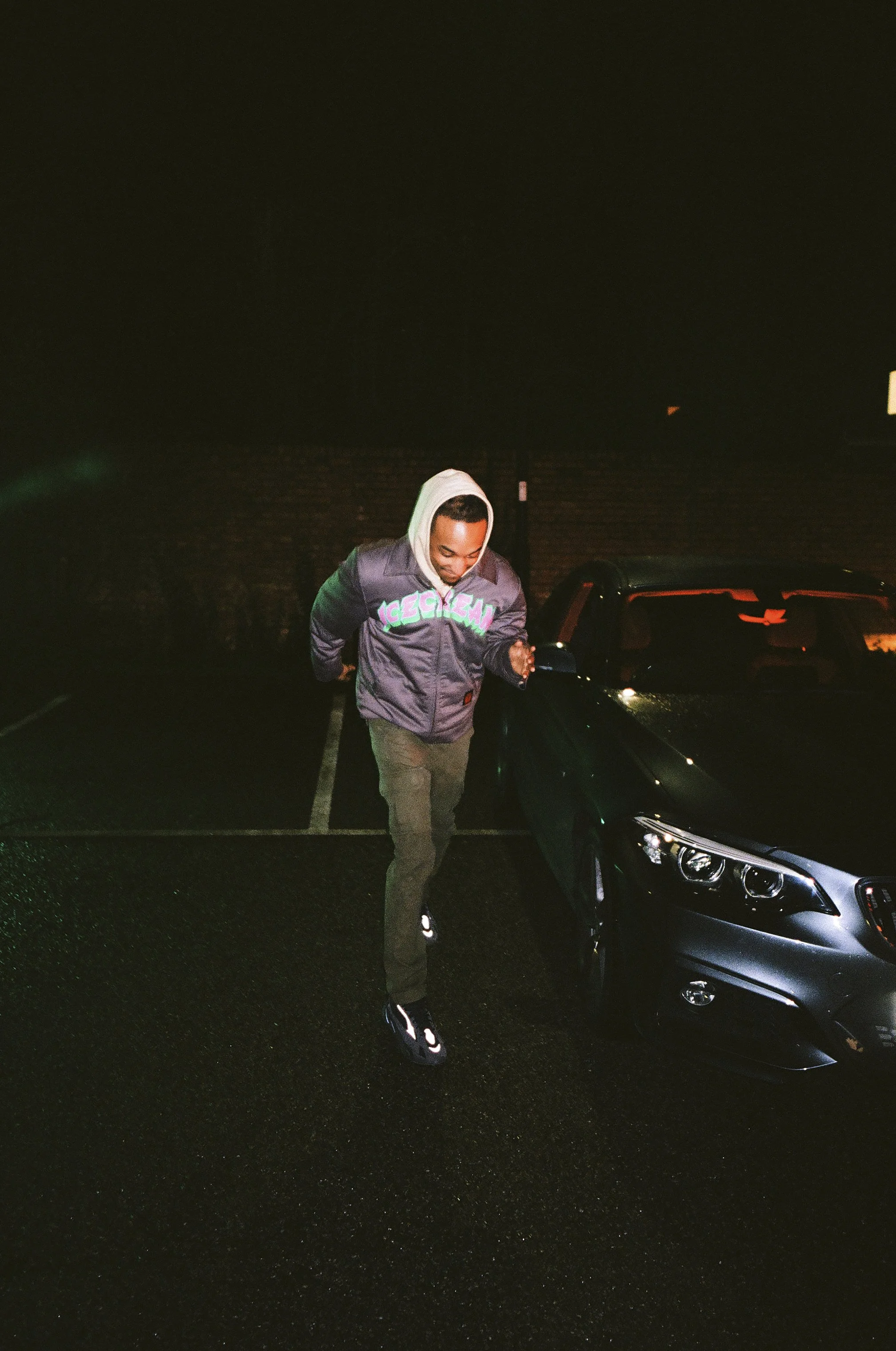 A man wearing a jacket and hoodie rising from a parking lot at night with a car behind him.